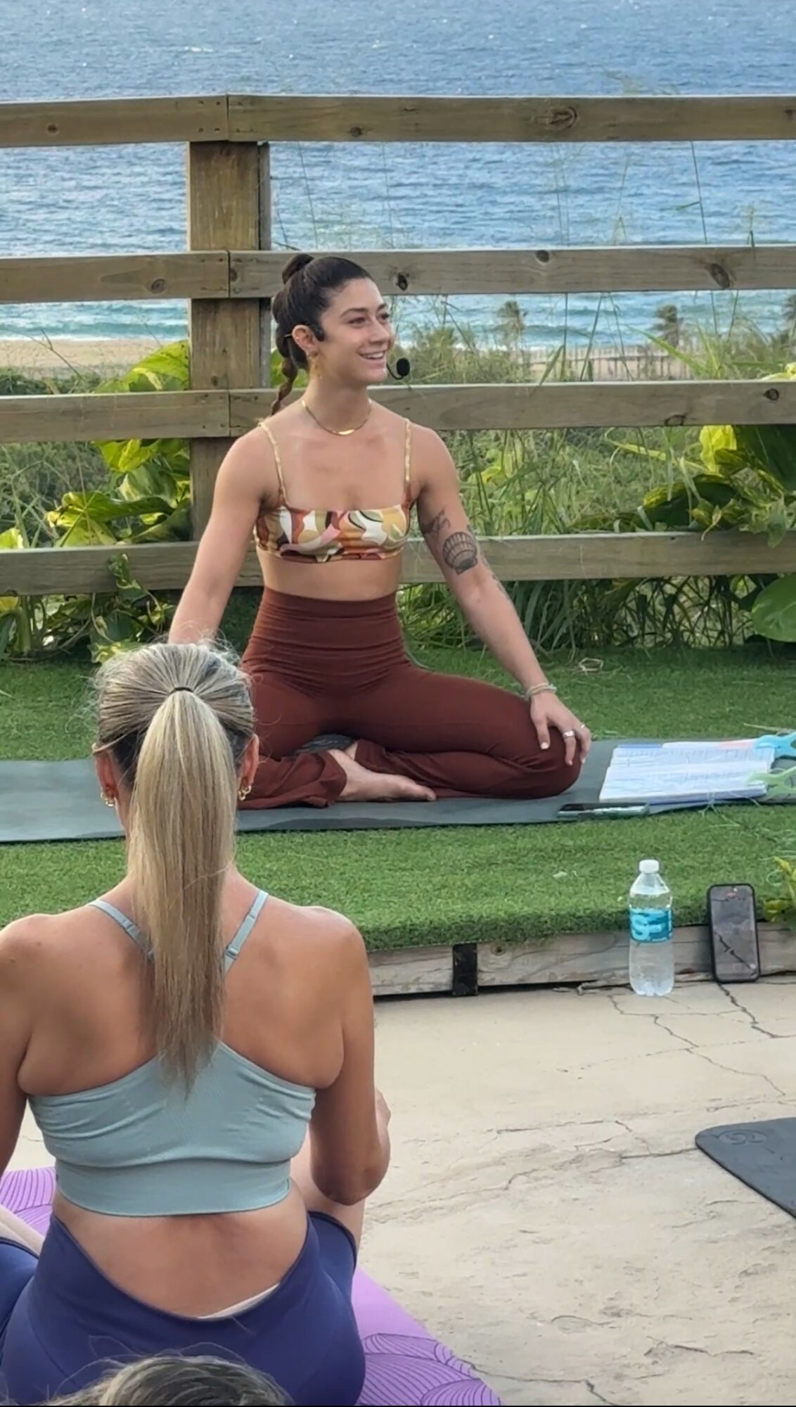 Woman practicing yoga outdoors on a mat with water and greenery in background, facing a seated person with blonde hair.