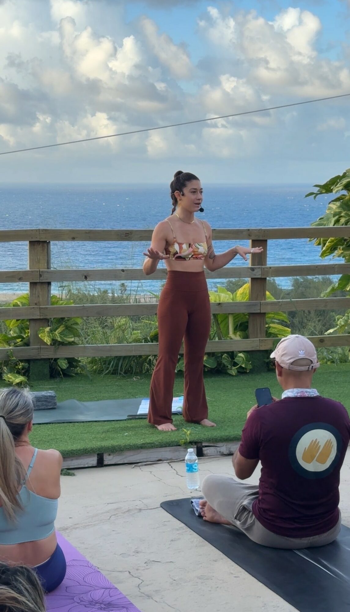 Woman standing on outdoor stage with ocean and sky background, holding microphone, wearing patterned top and high-waisted pants.