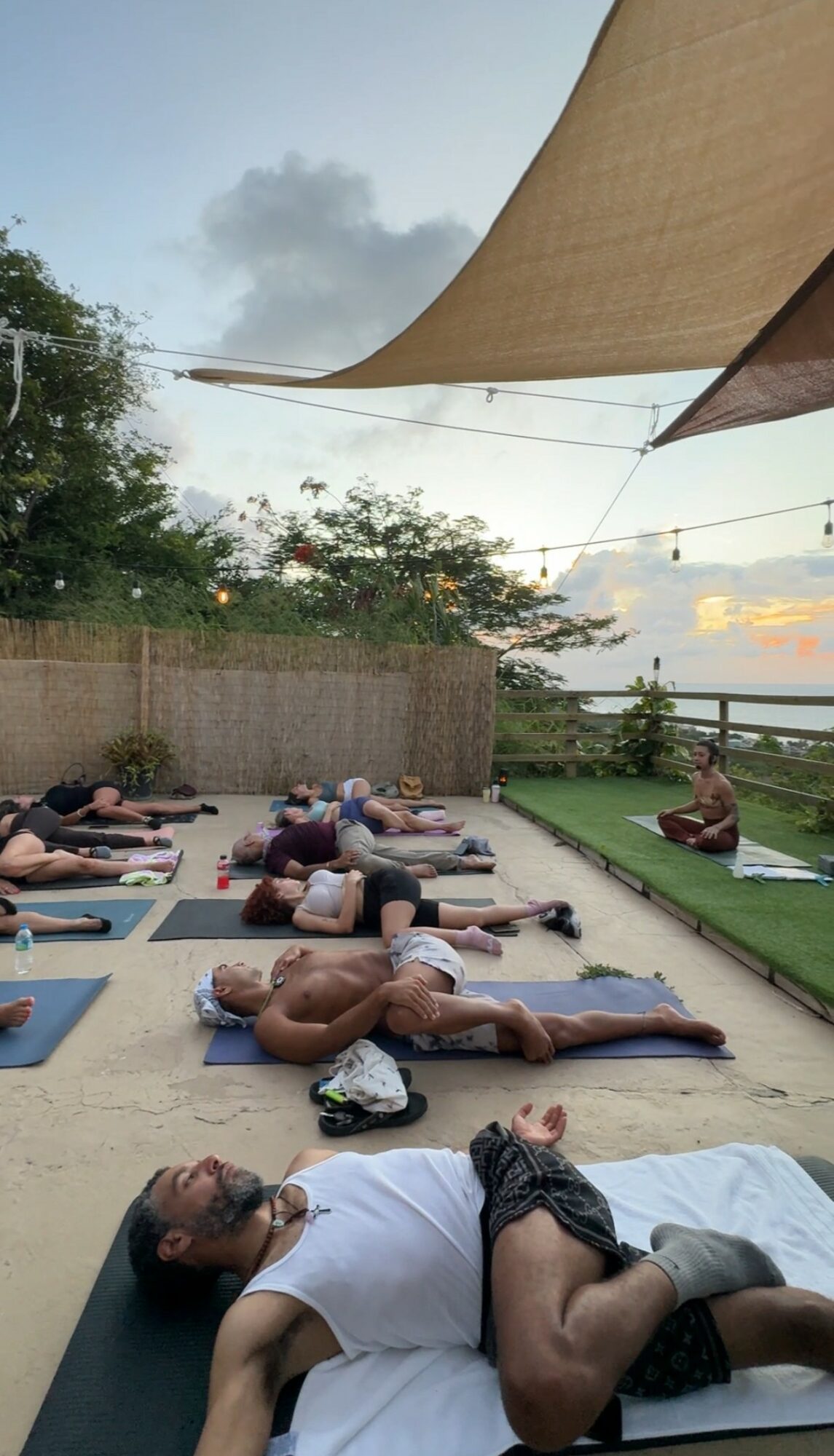 People practicing yoga outdoors on mats under a canopy, with trees and a fence in the background.
