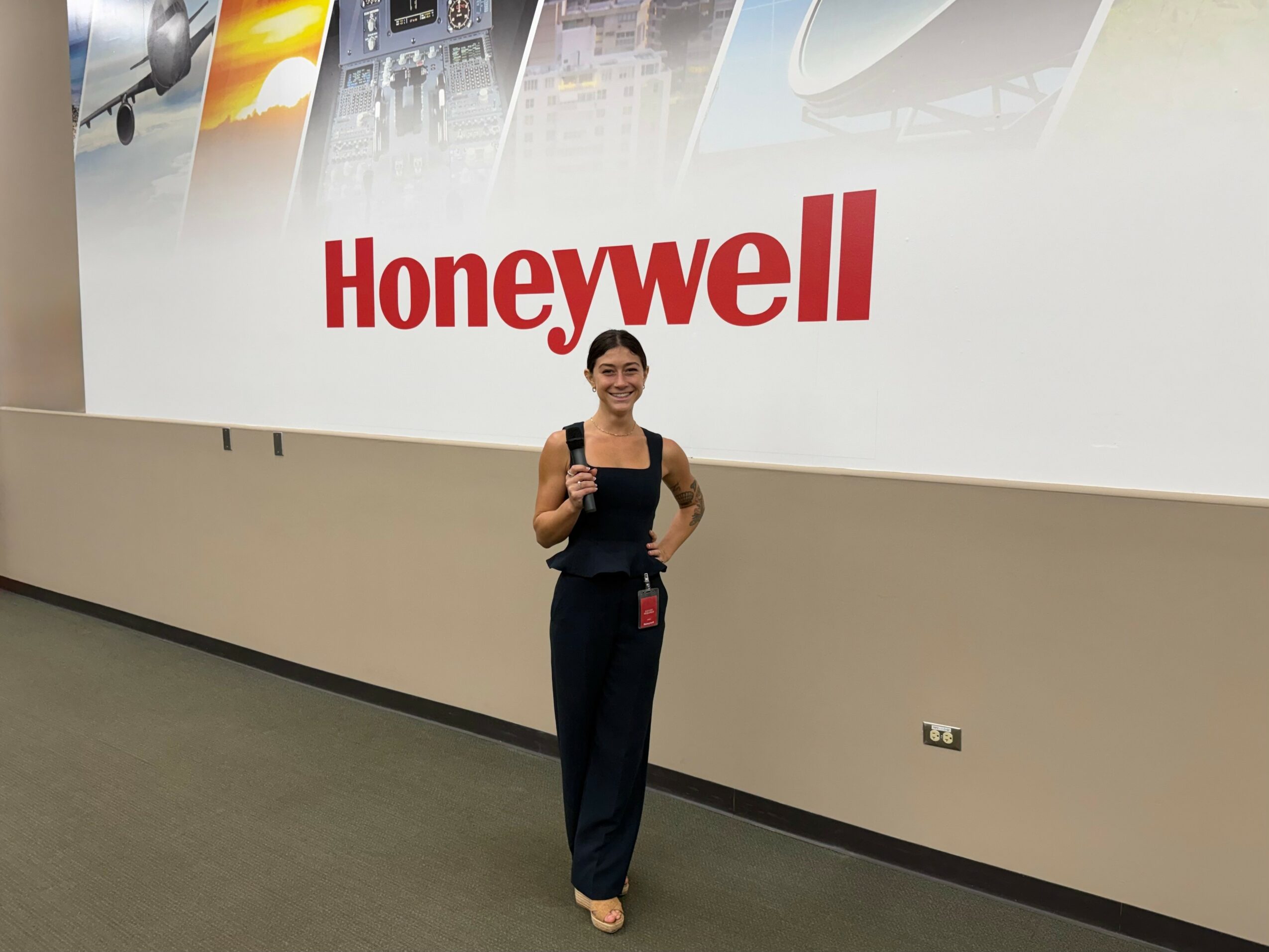 Woman standing in front of a Honeywell sign, smiling, with a backpack and badge, in an indoor setting.