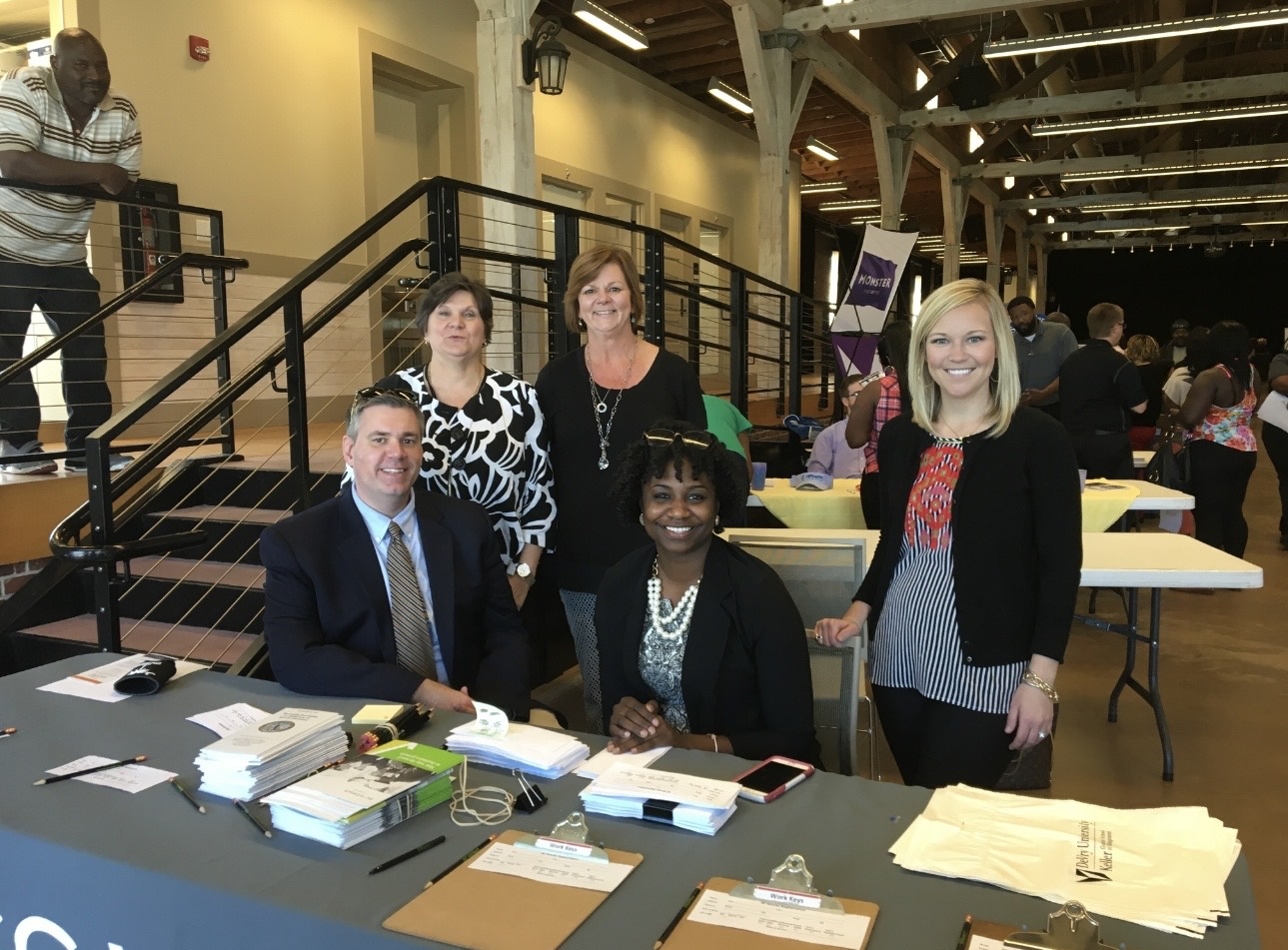 Group of five people standing behind a table with papers and a phone, in a large indoor space.