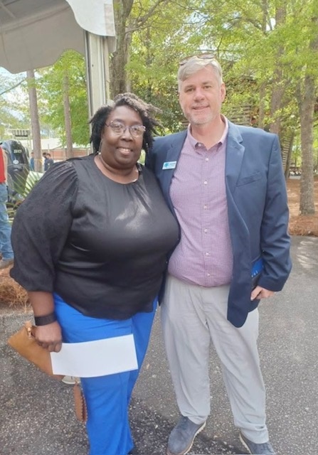 Two people standing outdoors in a wooded area, smiling, with trees and a parking lot in the background.