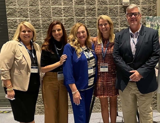 Group of five people standing together indoors, smiling, with a brick wall background.