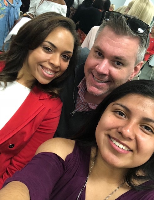 Three people smiling, taking a selfie indoors with other people in the background.