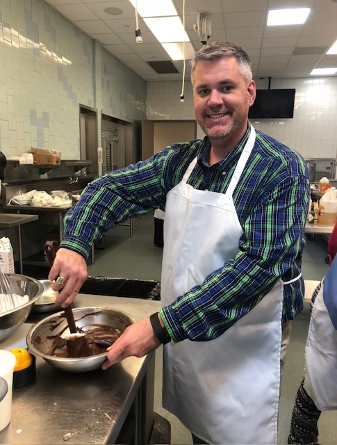 Man in plaid shirt and white apron stirring chocolate in a bowl in a kitchen.