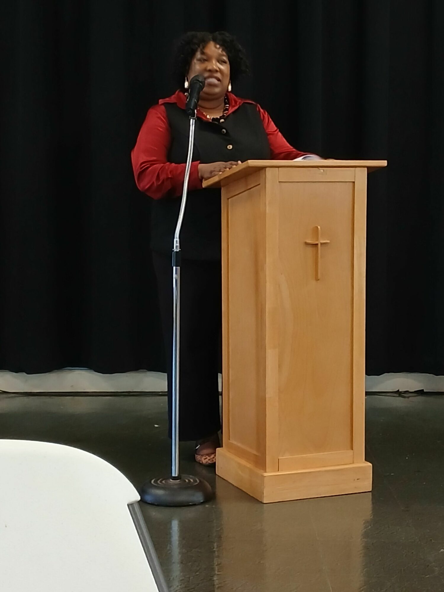 Woman standing at a wooden podium with a cross, speaking into a microphone on a stage with black curtains.