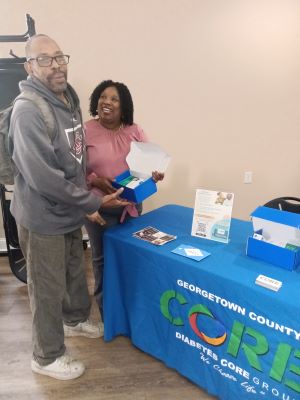 Two people standing at a table with informational materials, in a room with plain walls and wooden floor.