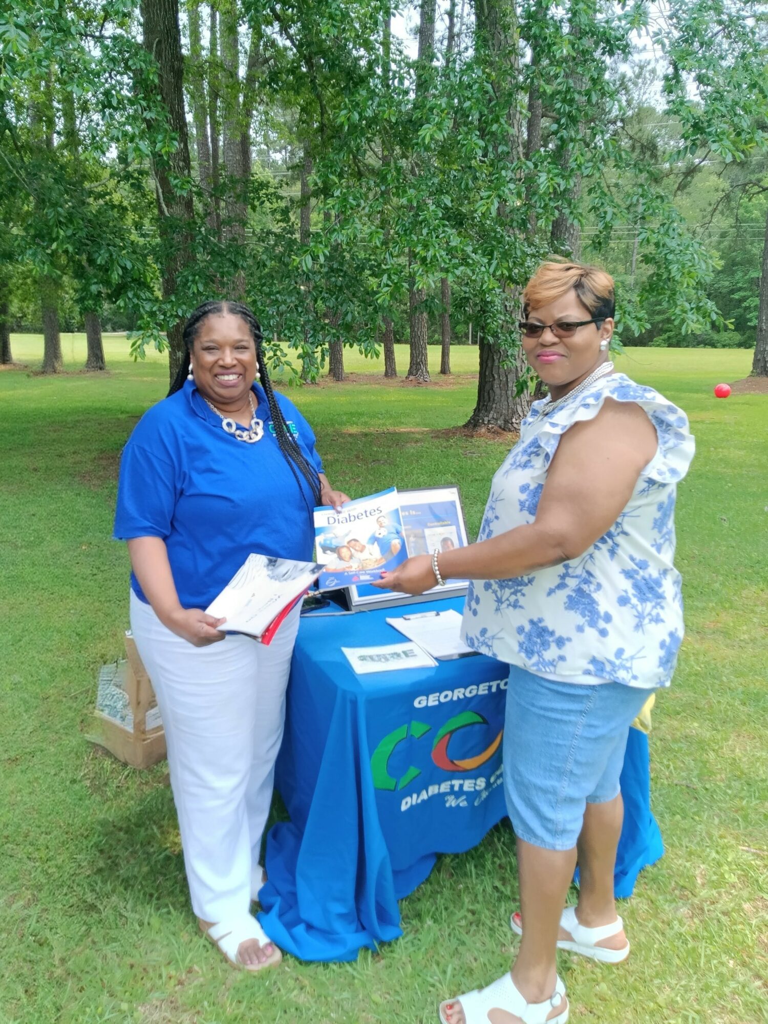 Two women standing outdoors near a table with a blue tablecloth, holding a book and smiling.