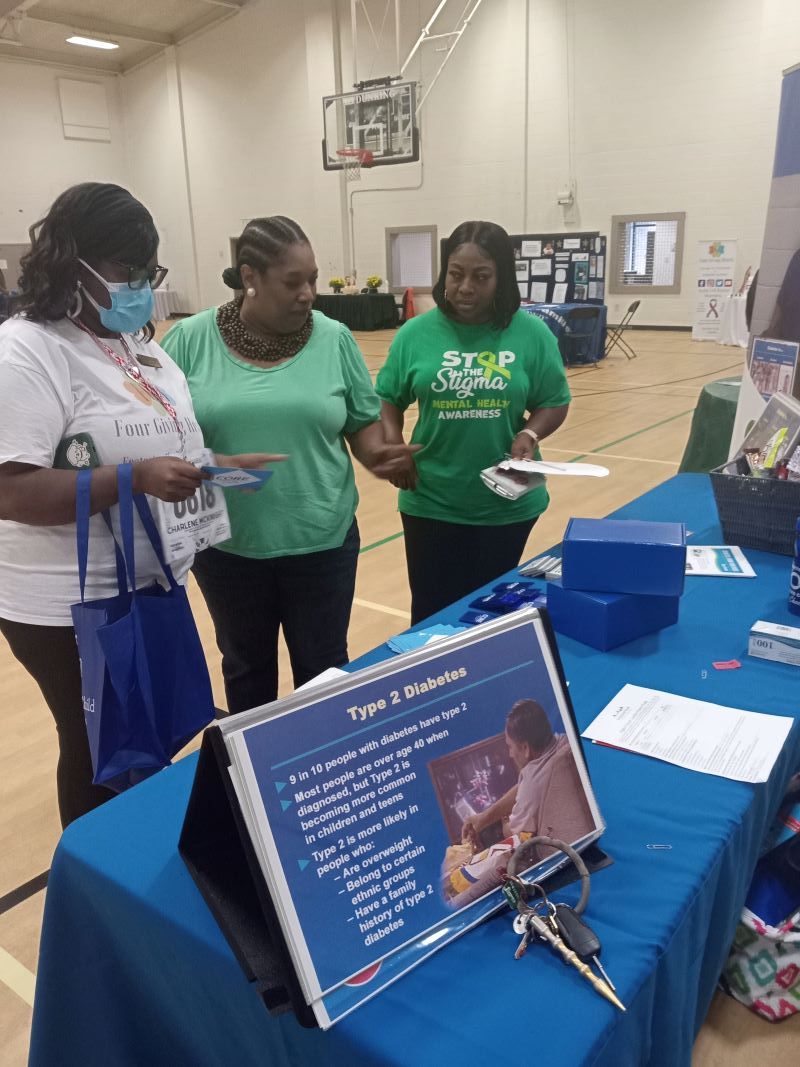 Four women in a gymnasium, two wearing green shirts, discussing health information at an informational booth with a blue tablecloth.