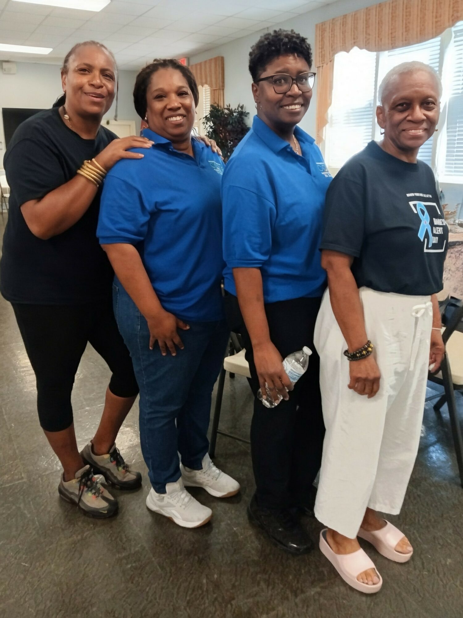 Four women standing in a row indoors, smiling, with curtains and chairs in the background.