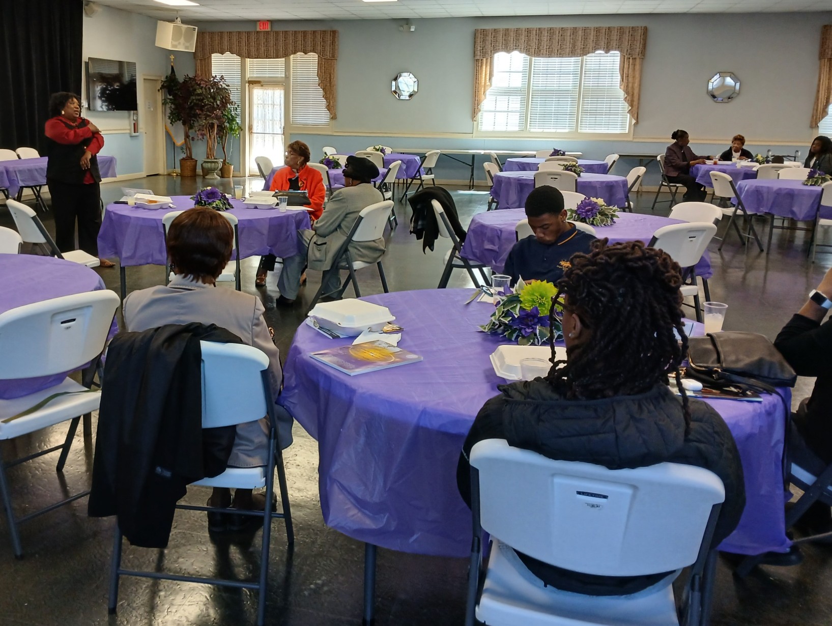 People seated at round tables with purple tablecloths in a well-lit room, some engaging in conversation, others using devices.