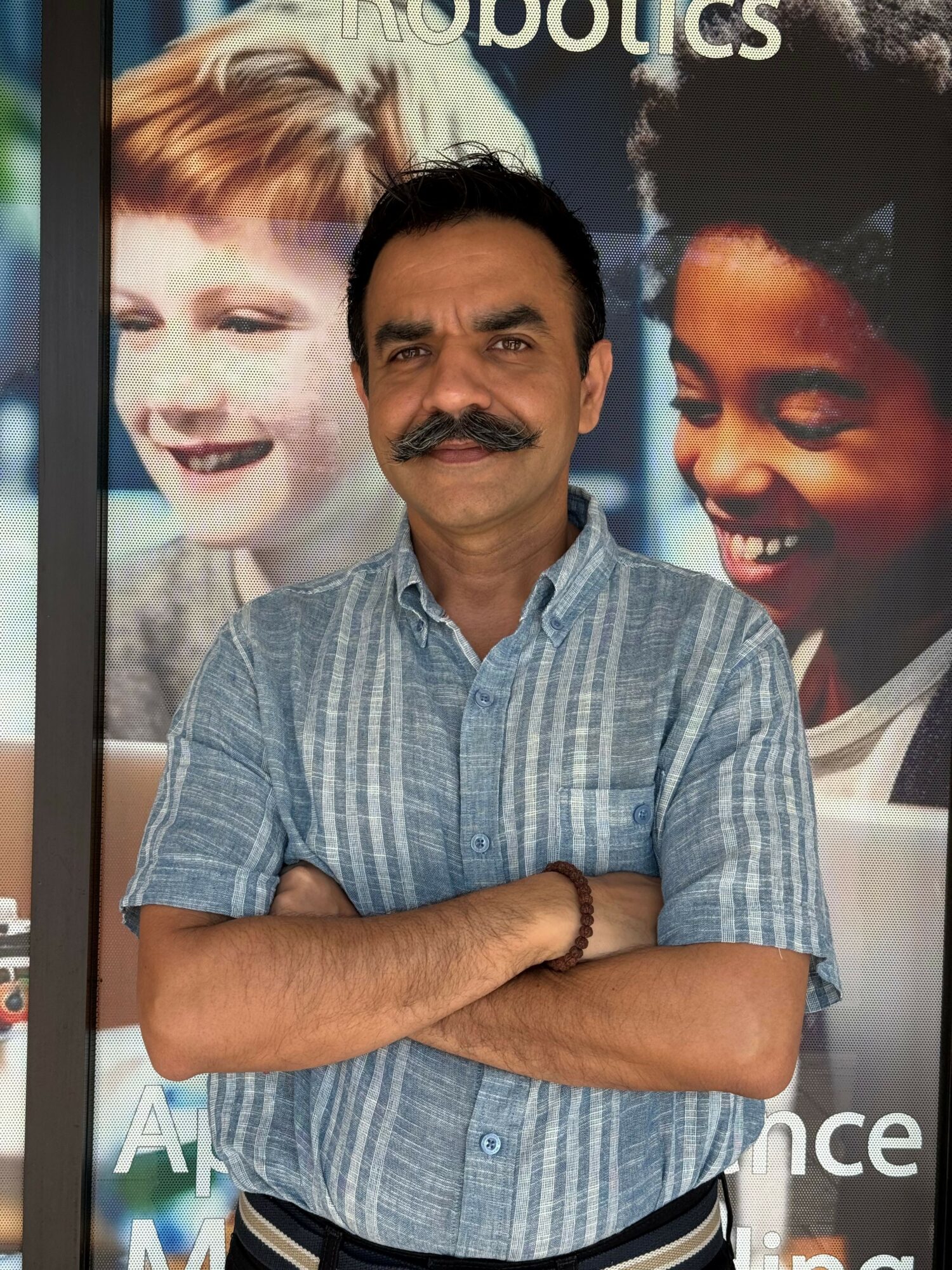 Man with mustache and short black hair standing with arms crossed in front of a poster with children.