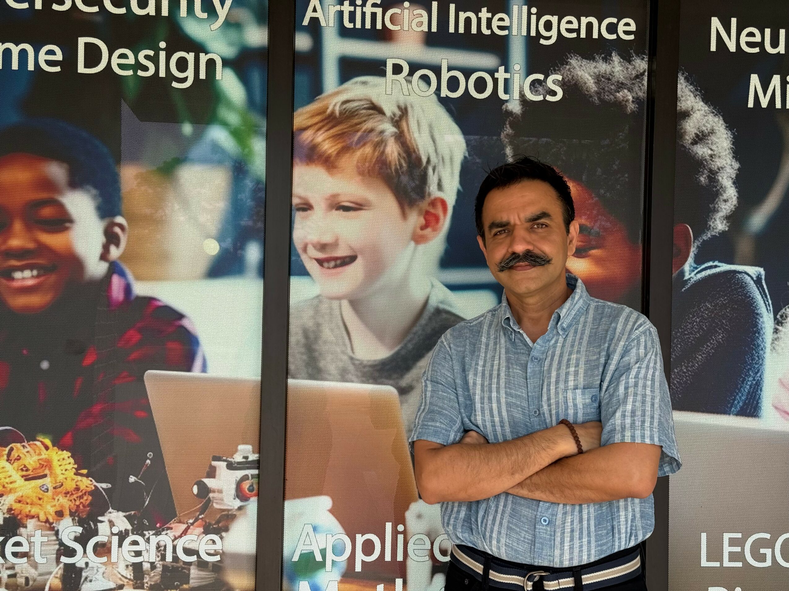 Man with mustache and striped shirt standing with arms crossed in front of large posters of children and technology.