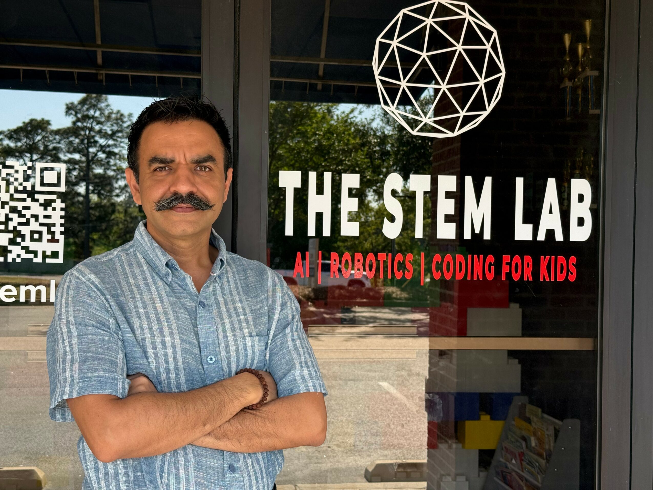 Man with mustache and crossed arms standing outside STEM lab with sign and geometric light fixture.