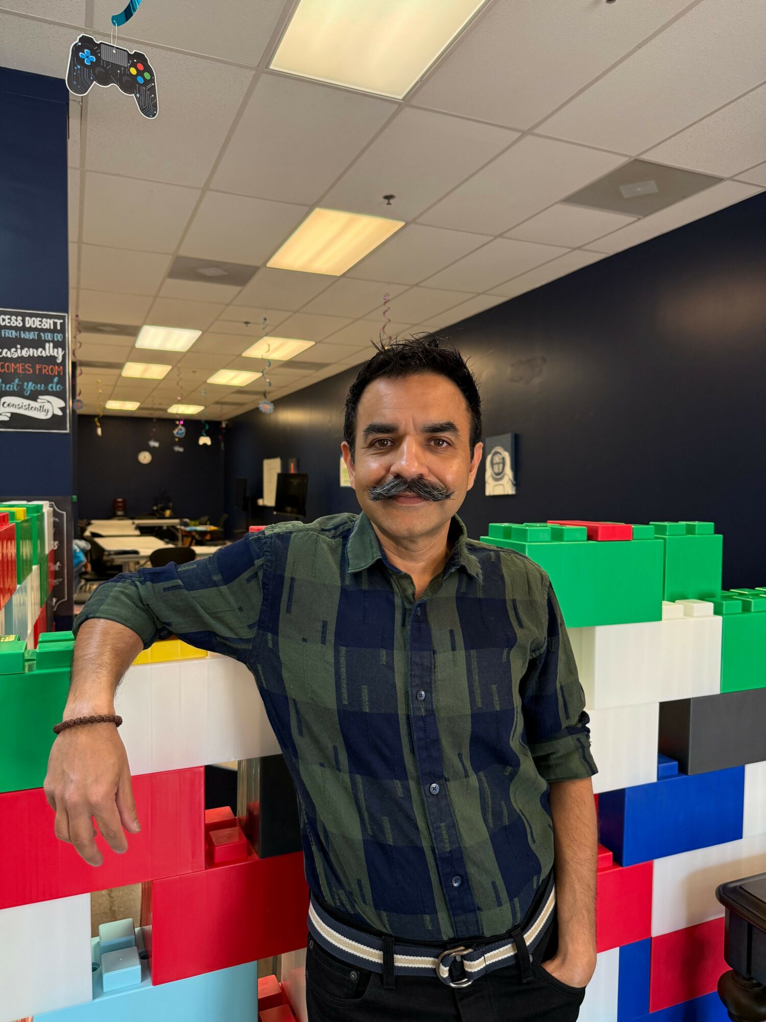 Man with mustache and dark hair in a green and black checkered shirt, standing indoors with colorful blocks behind him.