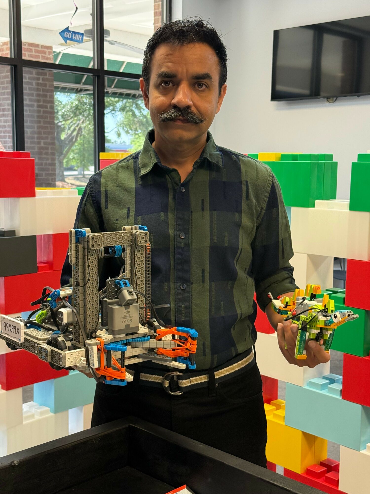 Man holding two LEGO creations, standing indoors with large windows and colorful LEGO structures in background.