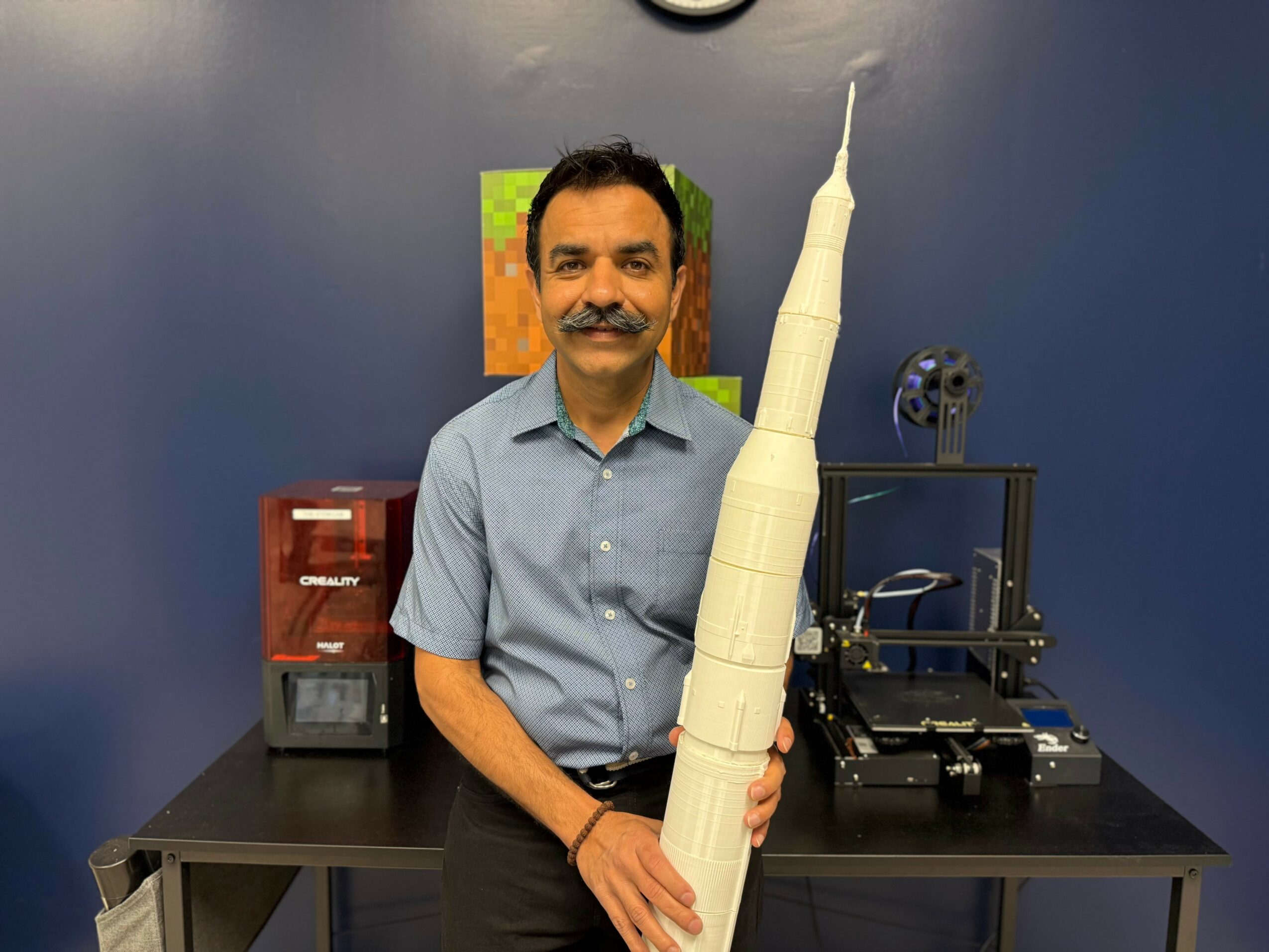 Man with a mustache in a short-sleeved shirt standing behind a table with a model of a tall building, blue wall background.