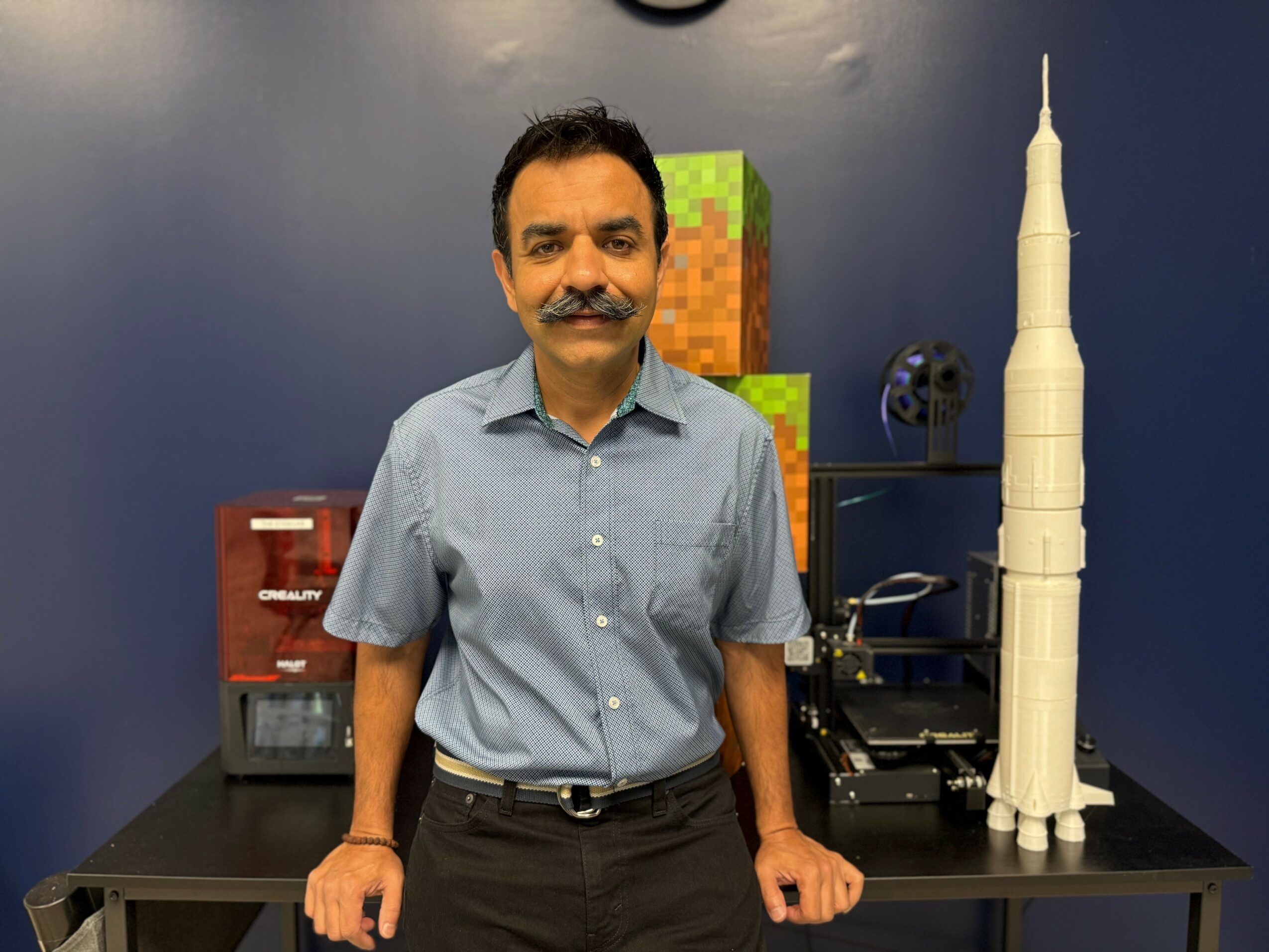 Man with mustache in light blue shirt standing behind a table with a model rocket and electronic equipment.