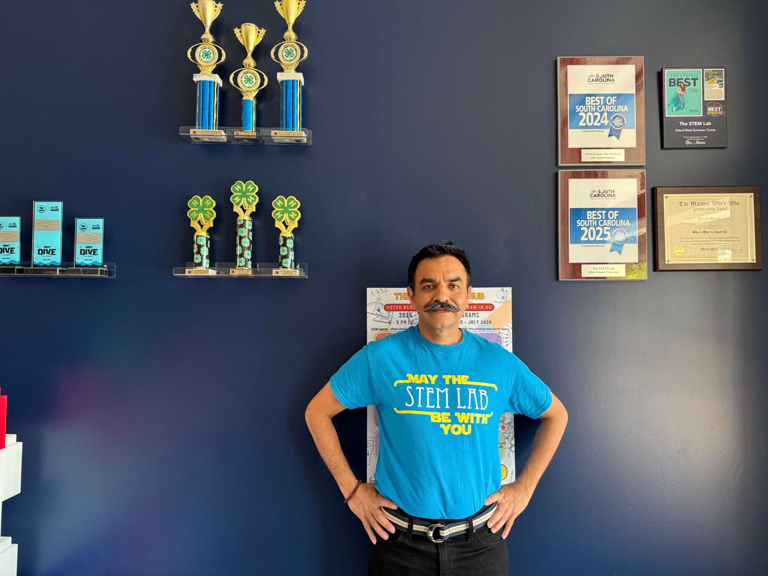 Man sitting in front of a dark blue wall with trophies, certificates, and awards displayed.