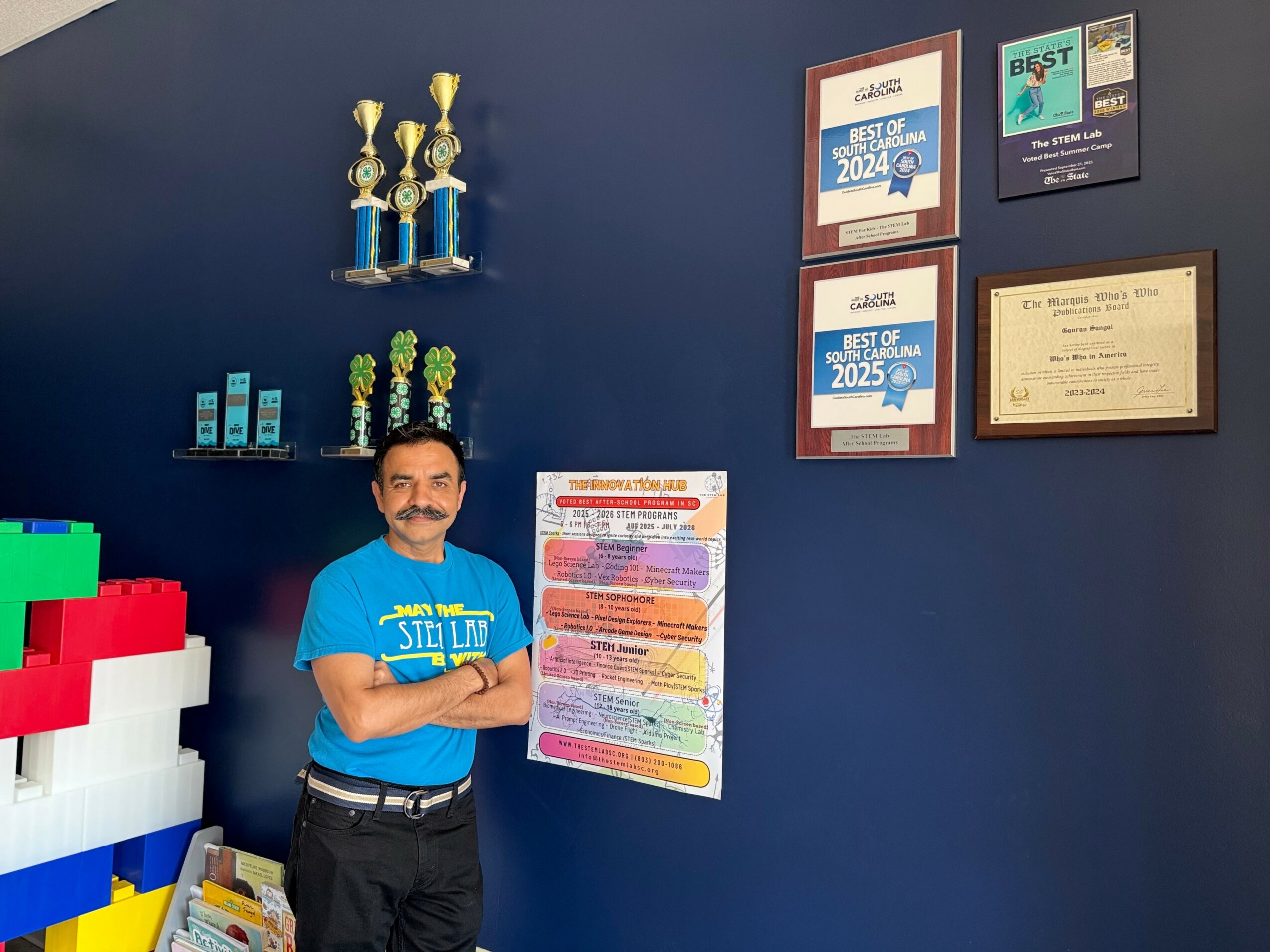 Man in blue shirt standing next to colorful poster on dark wall with awards and trophies displayed.