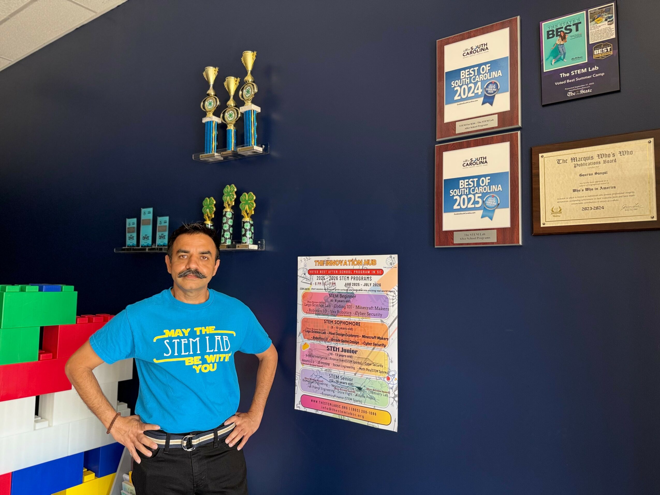 Man with a mustache and beard wearing a blue T-shirt standing indoors with awards and framed certificates on the wall behind him.