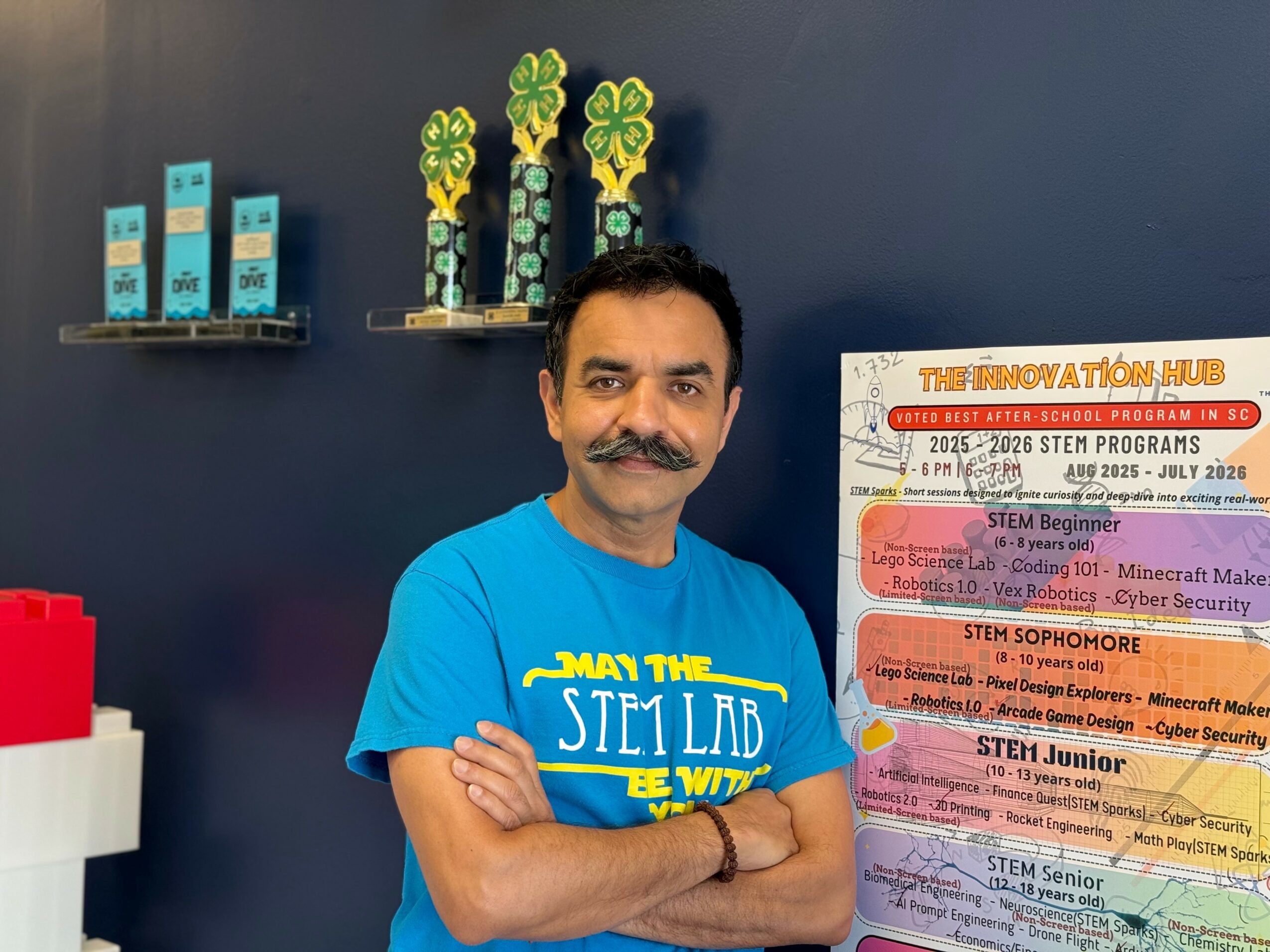Man with a mustache and blue T-shirt standing with arms crossed in front of a dark wall with shelves and a colorful poster.