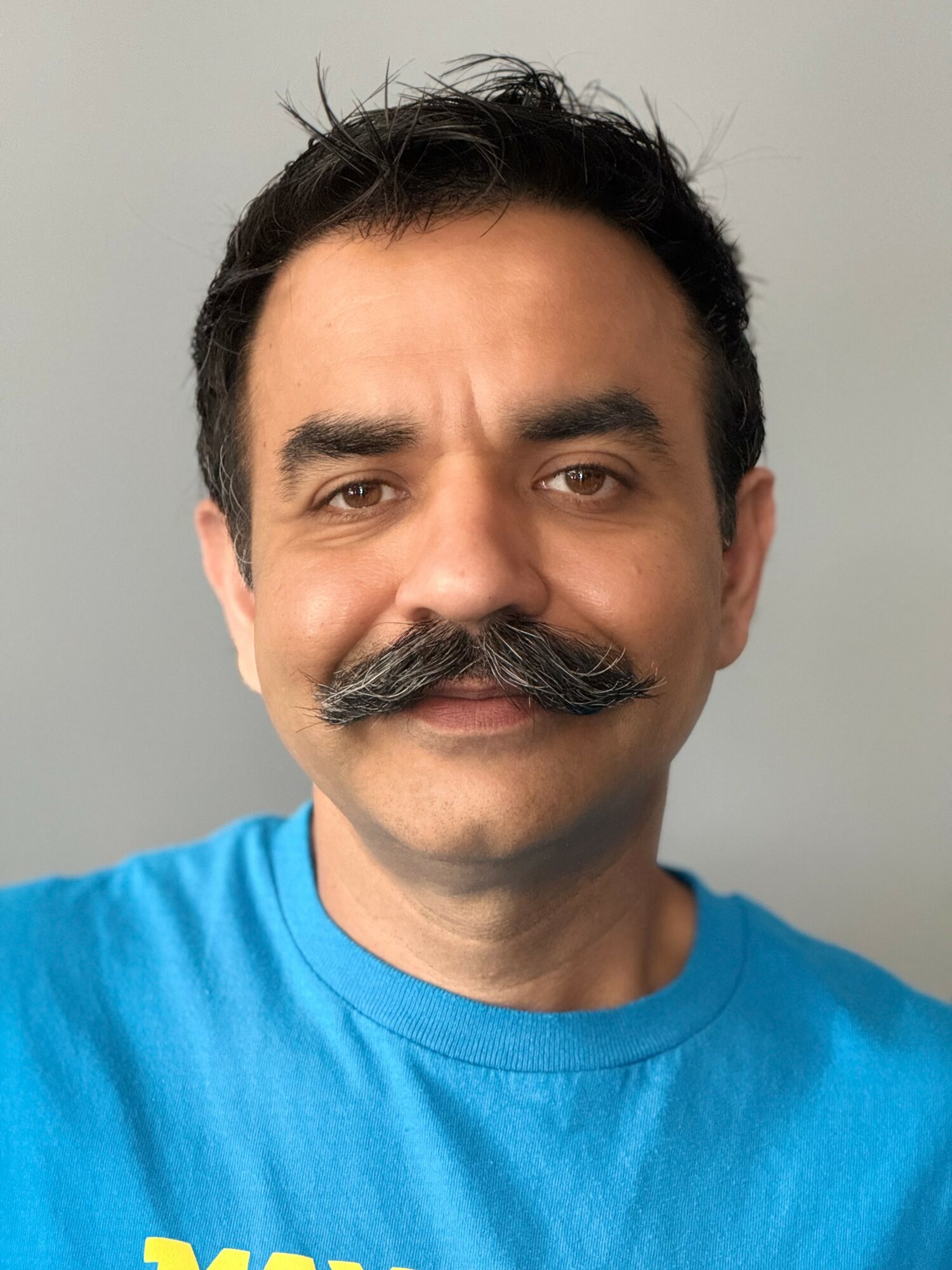 Man with dark hair and mustache wearing a blue shirt, smiling slightly, against a plain background.
