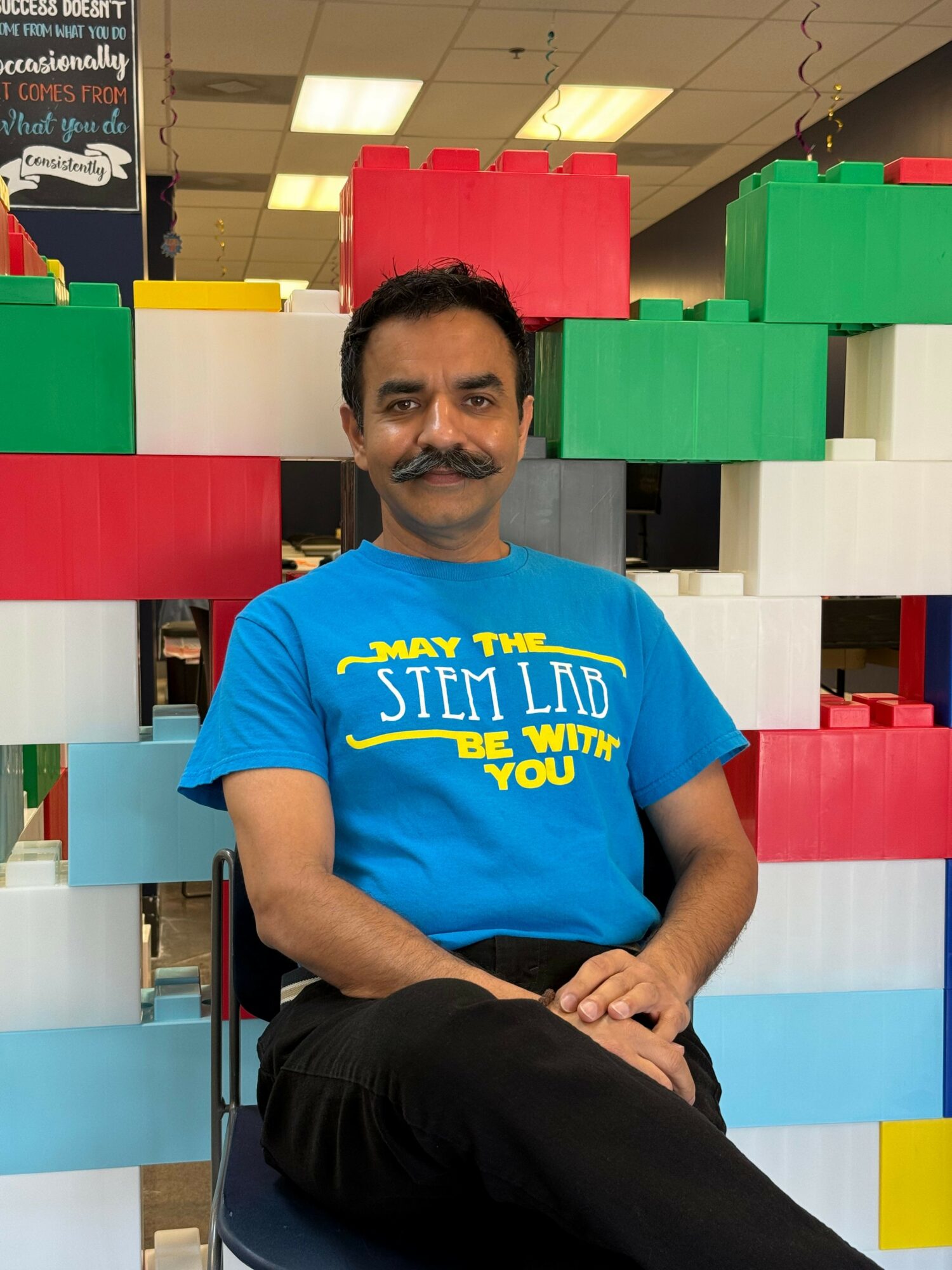 Man with mustache and blue shirt sitting in front of colorful large building blocks in indoor space.