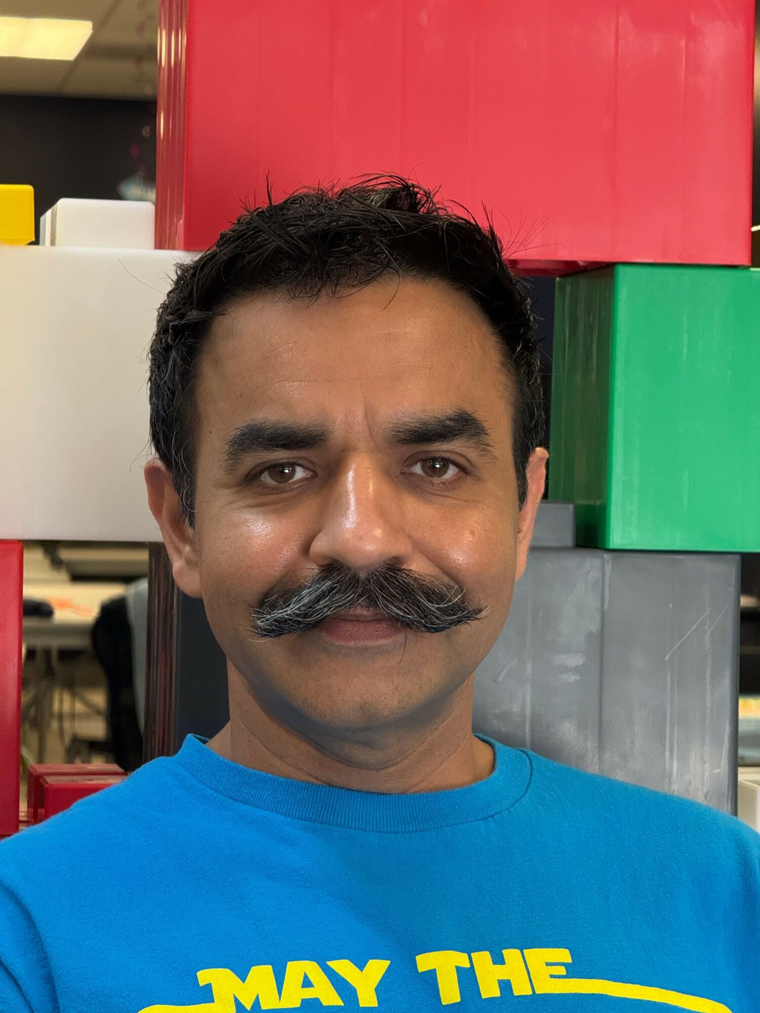 Man with dark hair and mustache wearing a blue shirt, standing indoors with colorful boxes behind him.