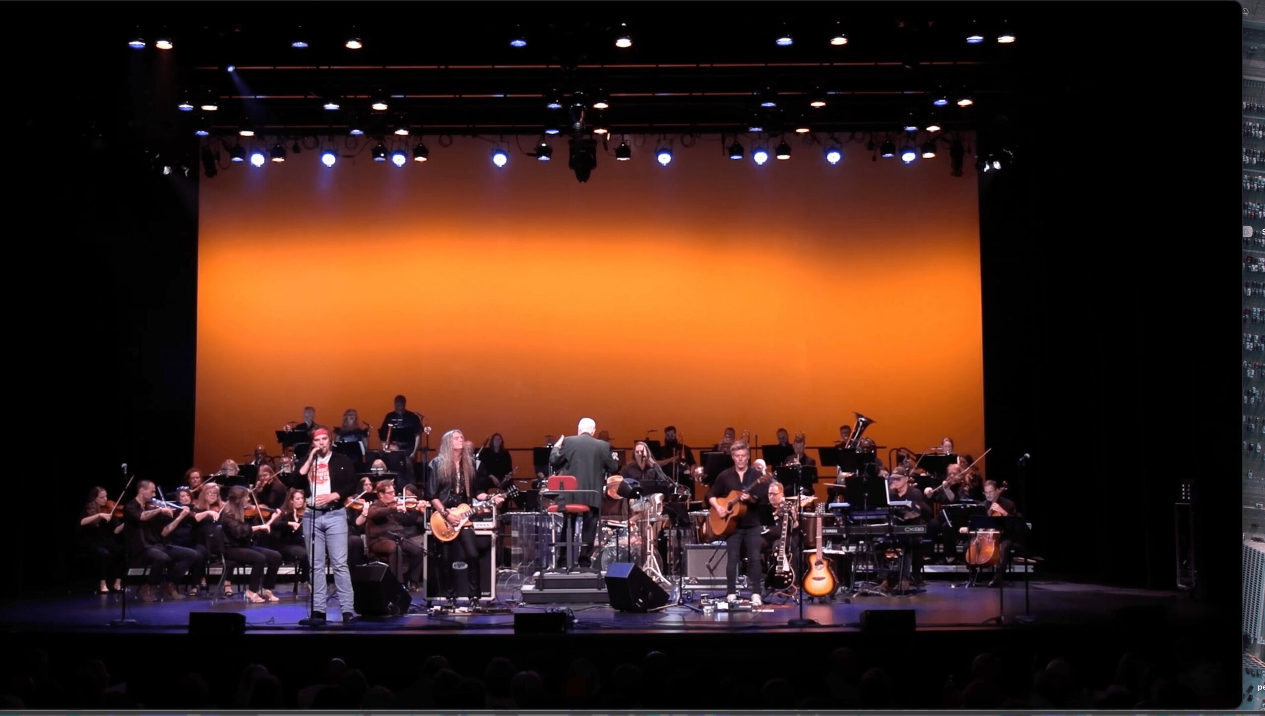 Orchestra on stage with conductor, musicians, and instruments, against a warm orange backdrop, in a concert hall.