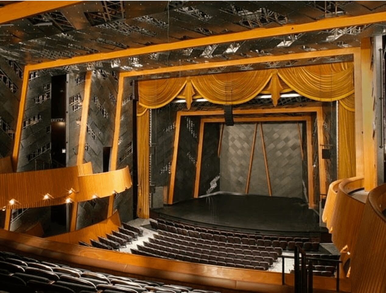 Empty theater stage with yellow curtains, black backdrop, and wooden framing, viewed from the audience area.
