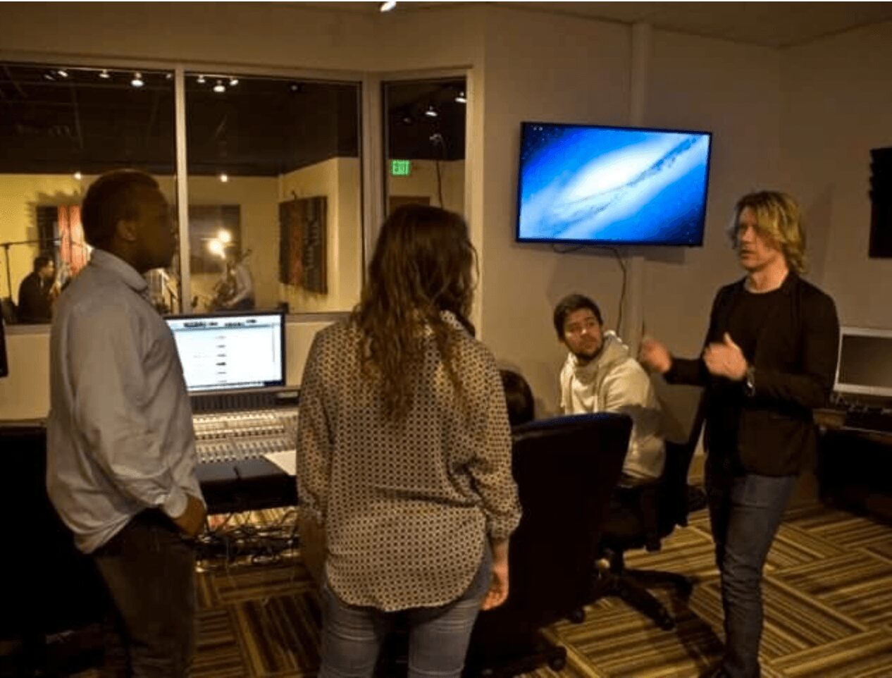 Group of five people in a music studio, one person speaking, others listening, computer and TV screen visible.