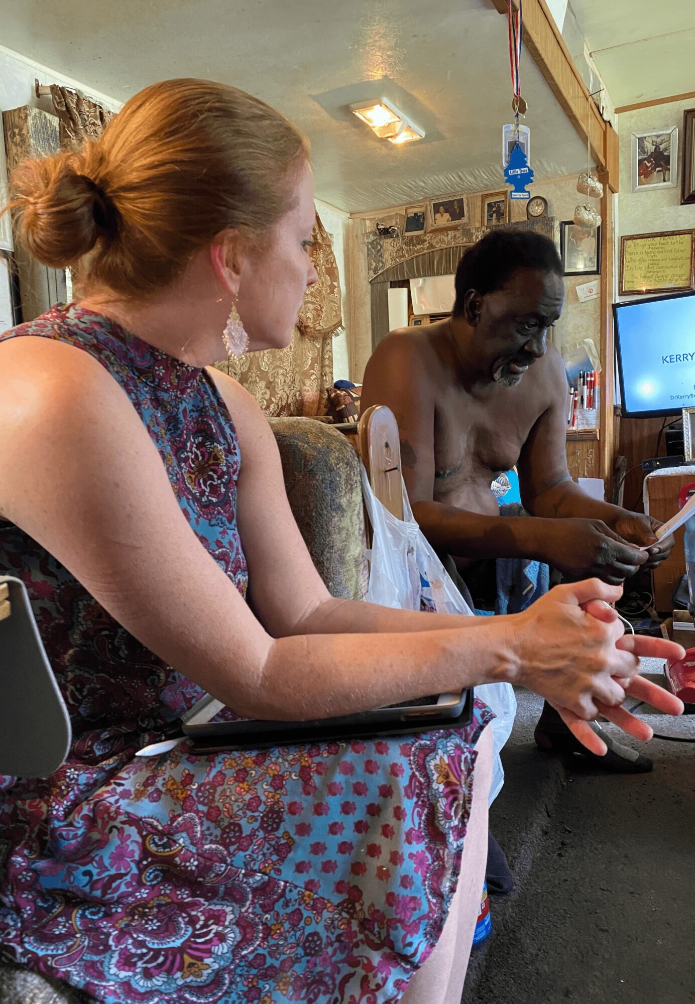 Two women sitting indoors, one with red hair in a bun and the other with dark skin, wearing a white dress.