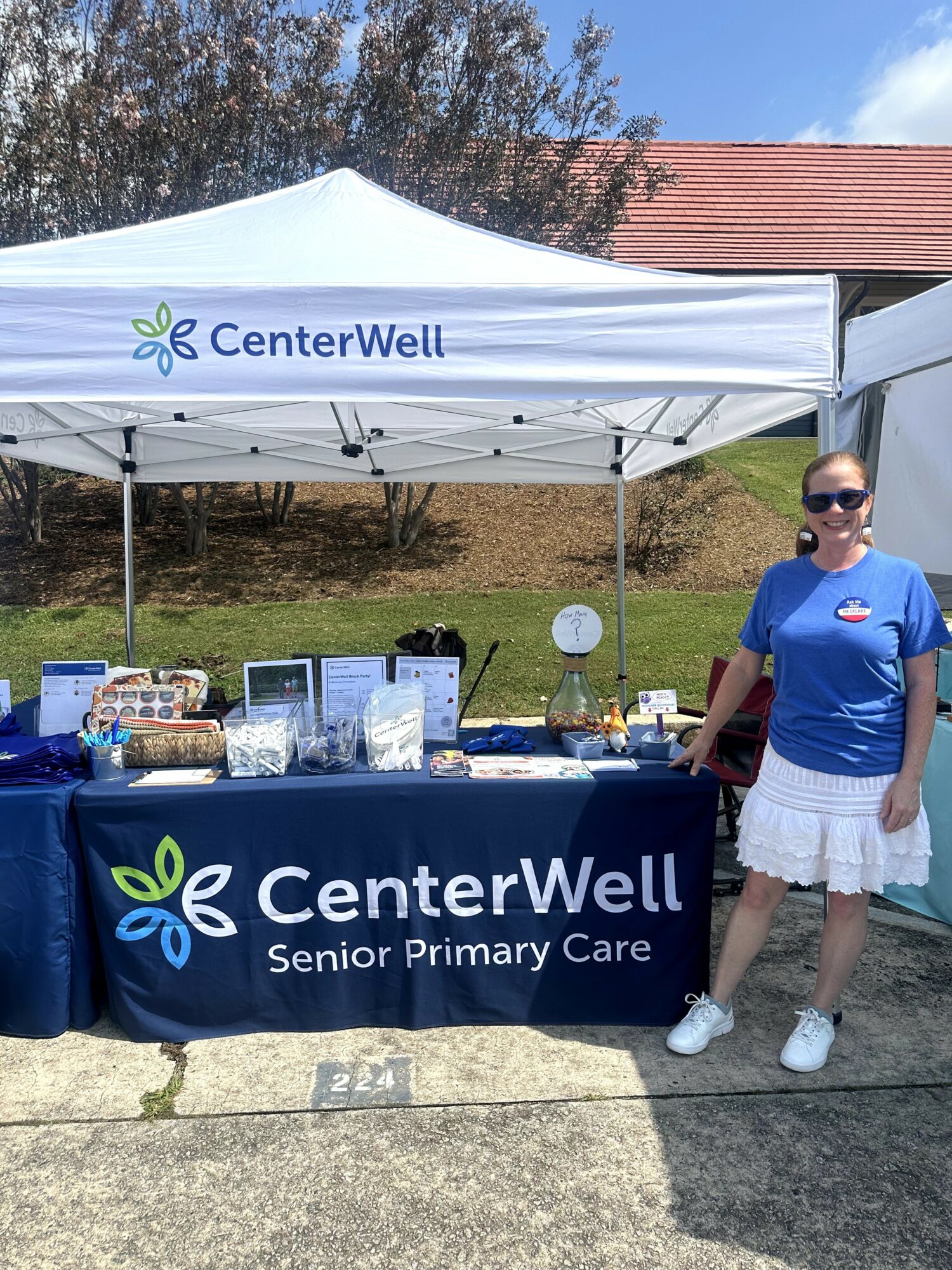 Woman in blue shirt and white skirt standing next to a table with CenterWell Senior Primary Care banner and items outdoors.
