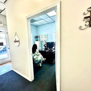 Office room with a person sitting at a desk and another person on a chair, visible through an open doorway.