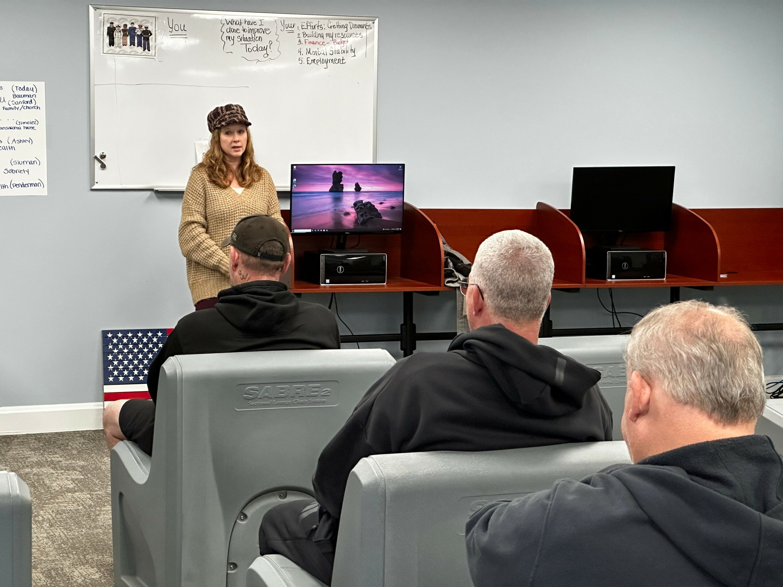 Woman presenting in front of a classroom with three men seated and listening.