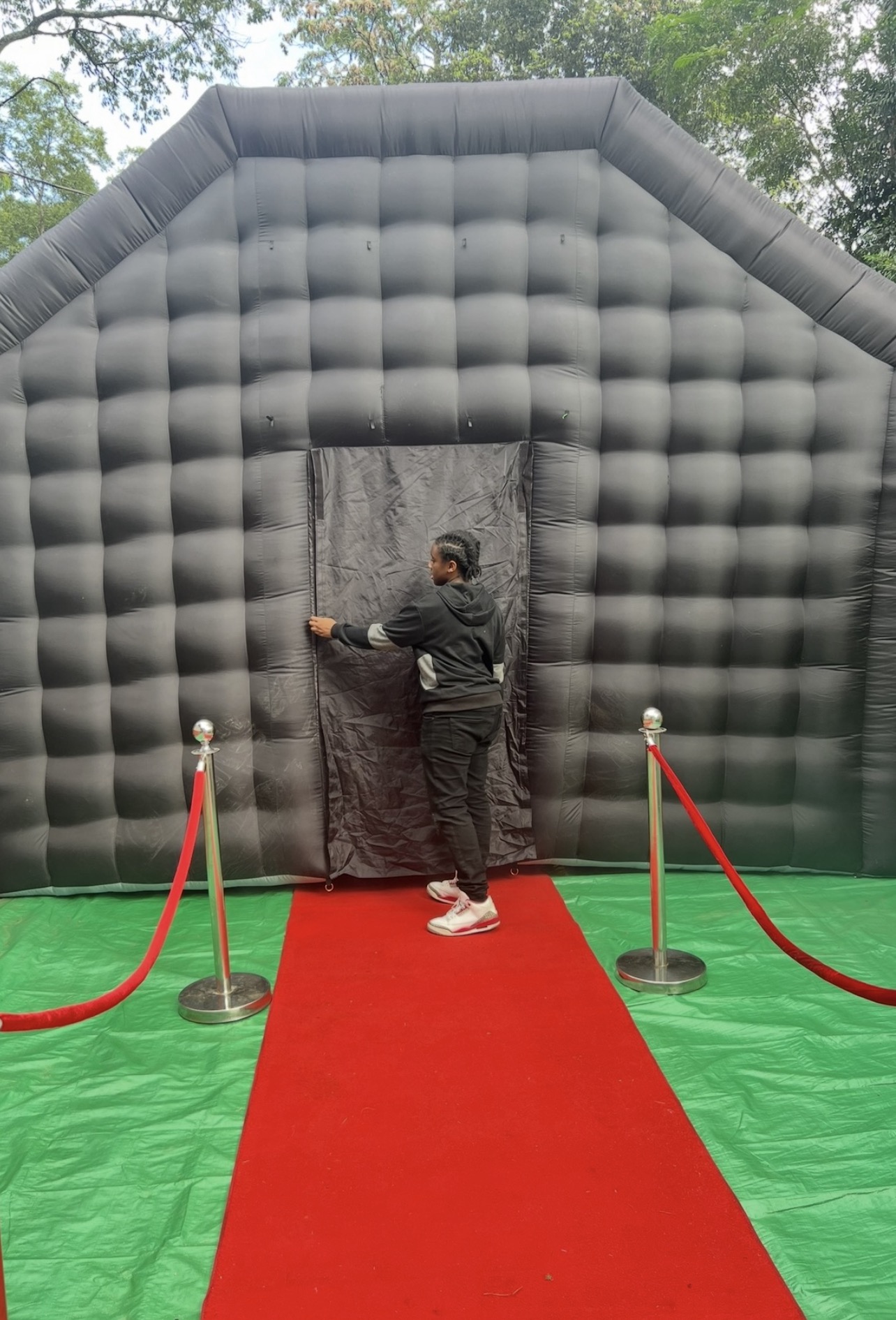 Person standing on red carpet in front of black inflatable structure with entrance, outdoors with trees in background.