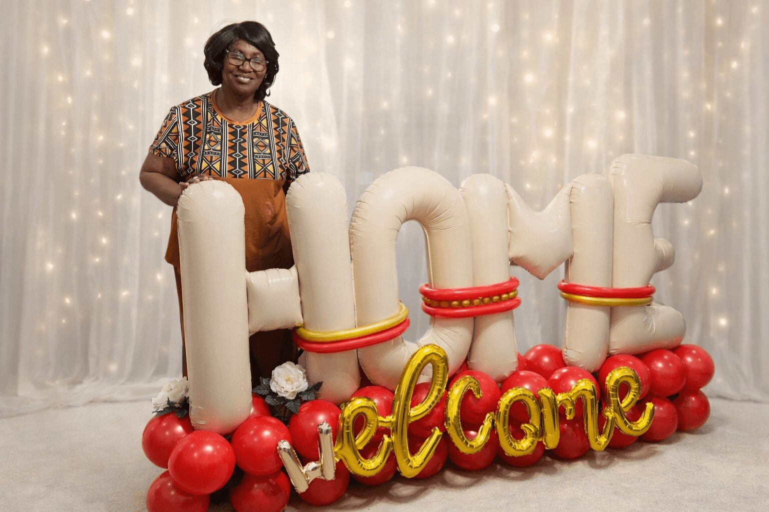 Woman standing behind large decorative letters spelling 'HOME' with a 'Welcome' sign at the bottom, in front of a fairy light backdrop.