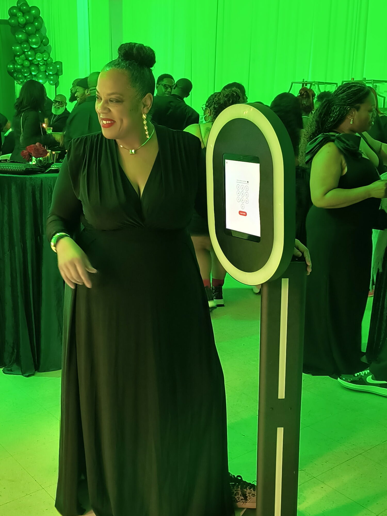 Woman in black dress smiling at event with green lighting and people in background.