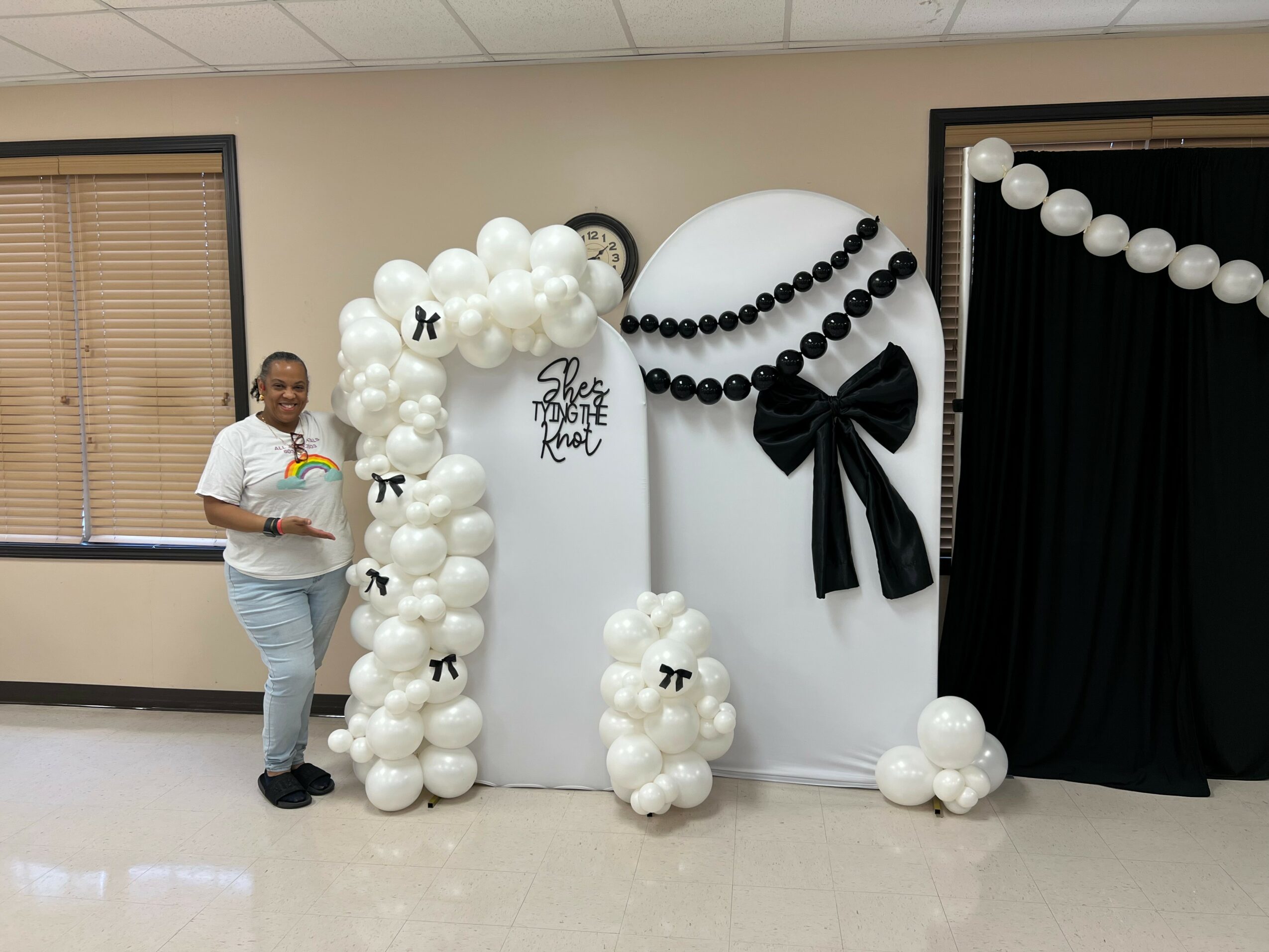 Person standing next to balloon decorations with black and white theme, including large balloons, bows, and a backdrop curtain.