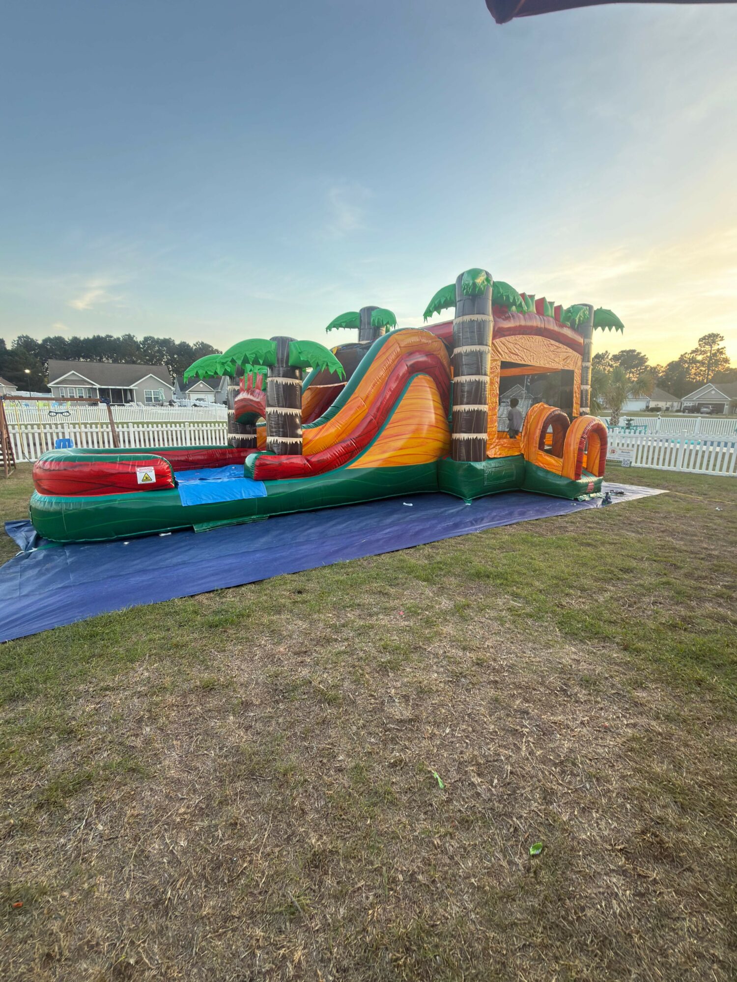 Colorful inflatable bounce house with slides and towers on grass, surrounded by white fence, under a clear sky.