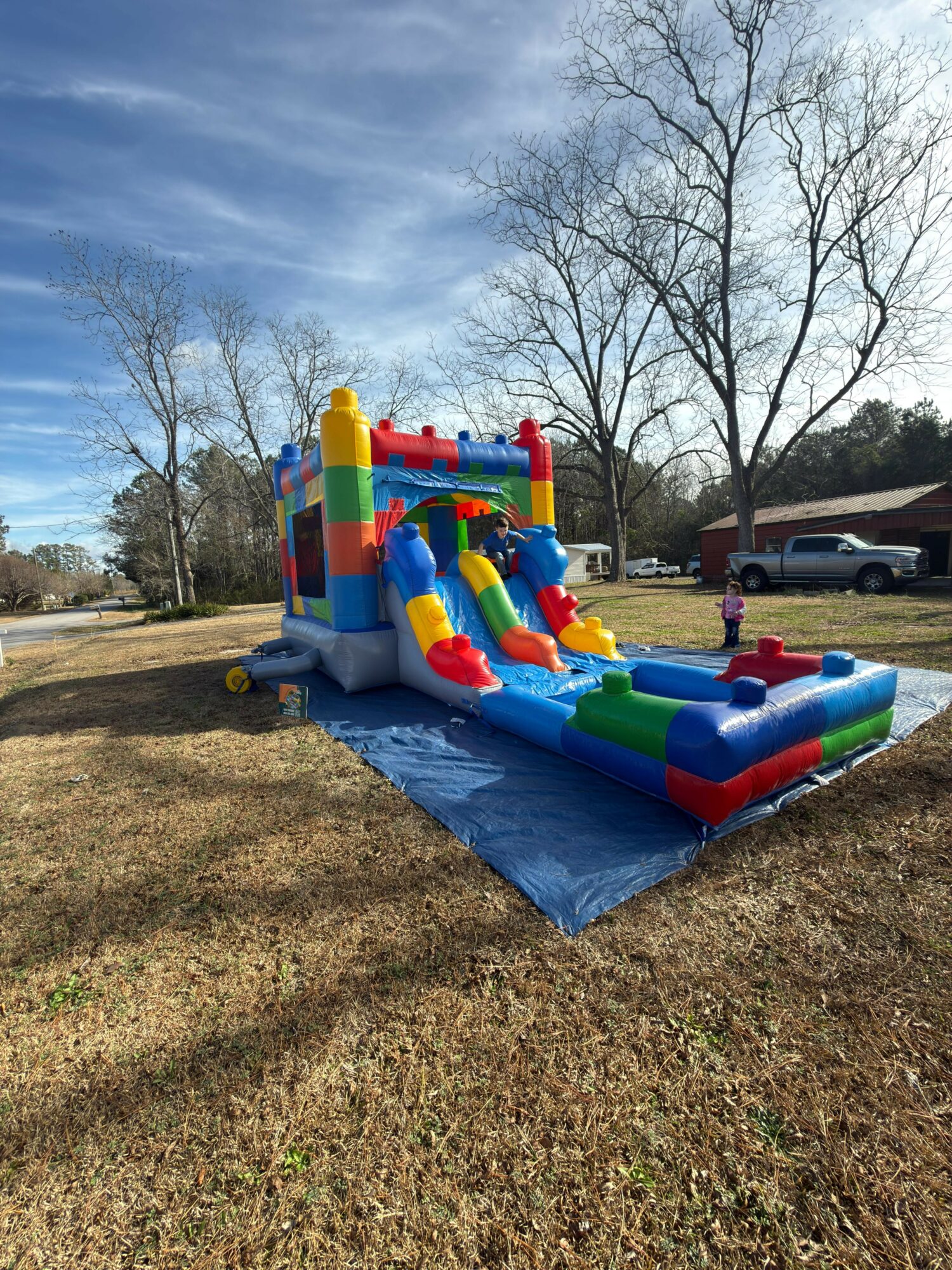 Colorful inflatable water slide with multiple slides and a splash area, set up outdoors on a blue tarp, with trees and cars in background.