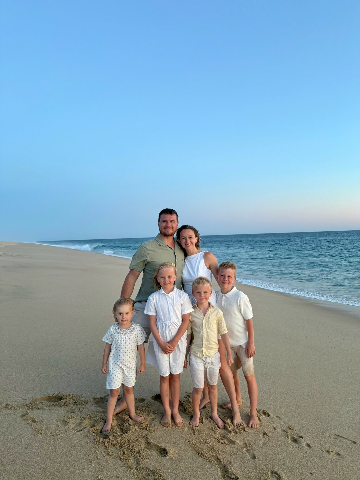 Group of seven people, children and adults, standing on a sandy beach near the ocean under a clear blue sky.