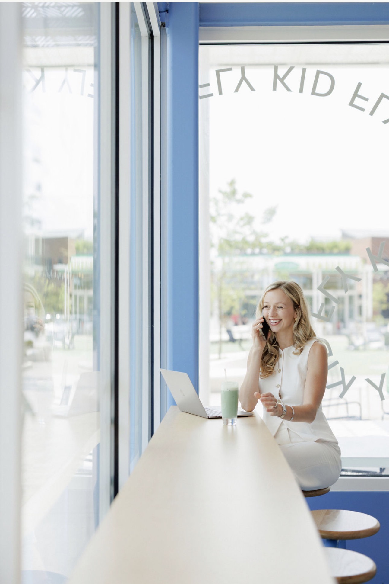 Woman sitting at a counter with a laptop and drink, smiling near large windows with outdoor view.