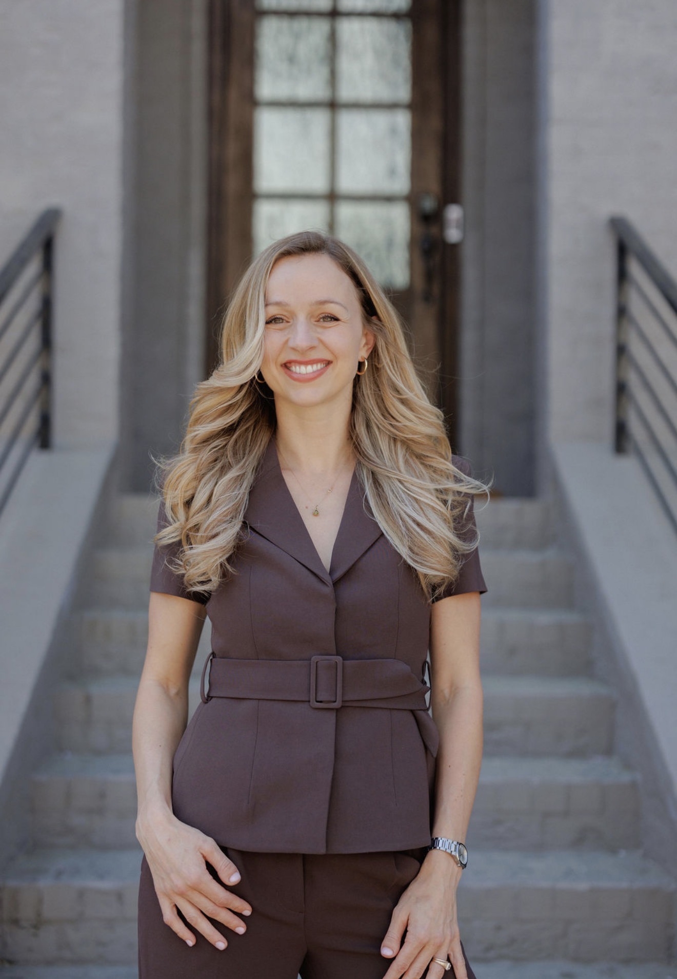 Woman with long wavy hair smiling, standing outdoors on stairs in front of a building door.