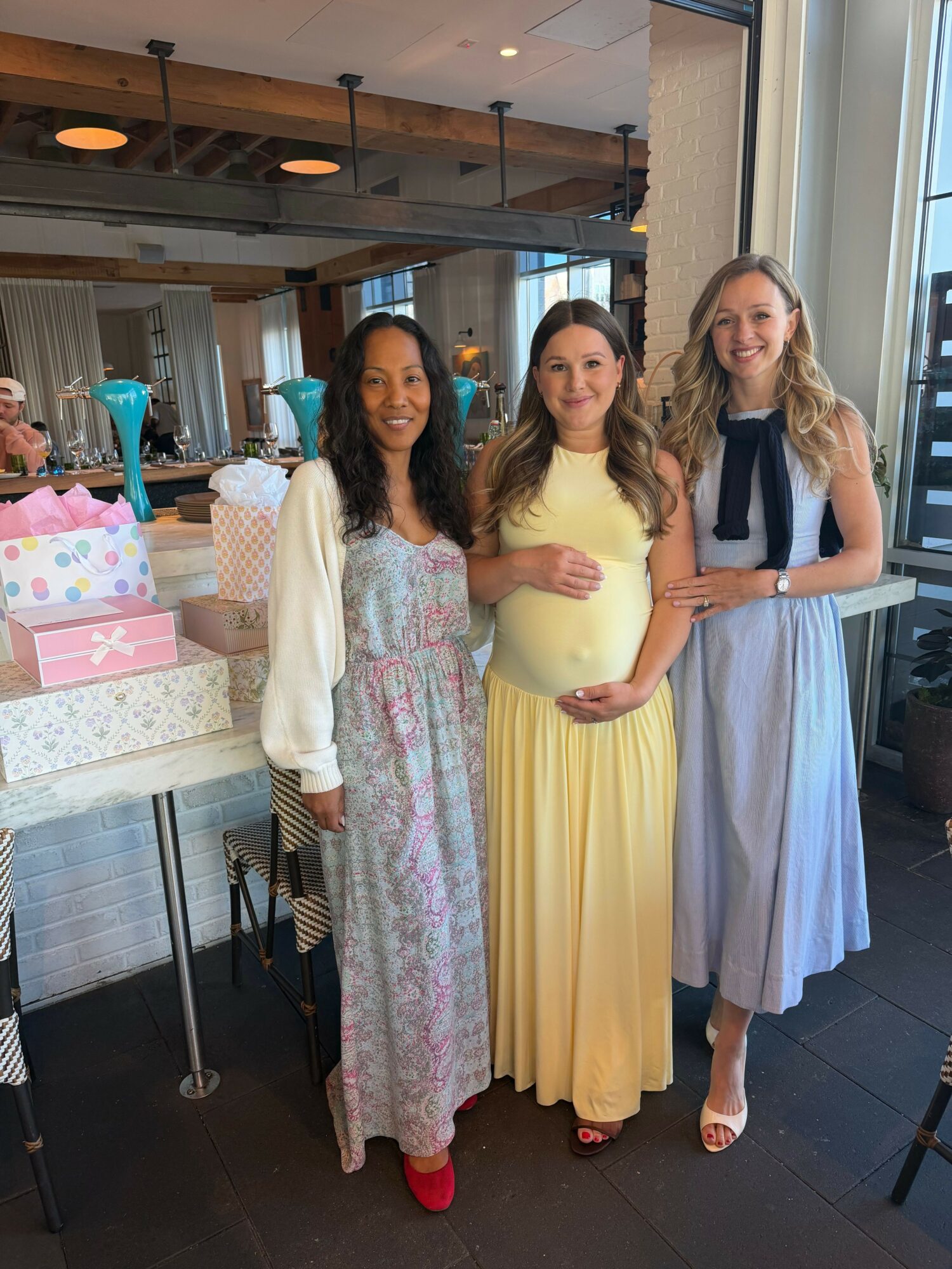 Three women standing indoors near a window, smiling, with gift boxes and tables in the background.
