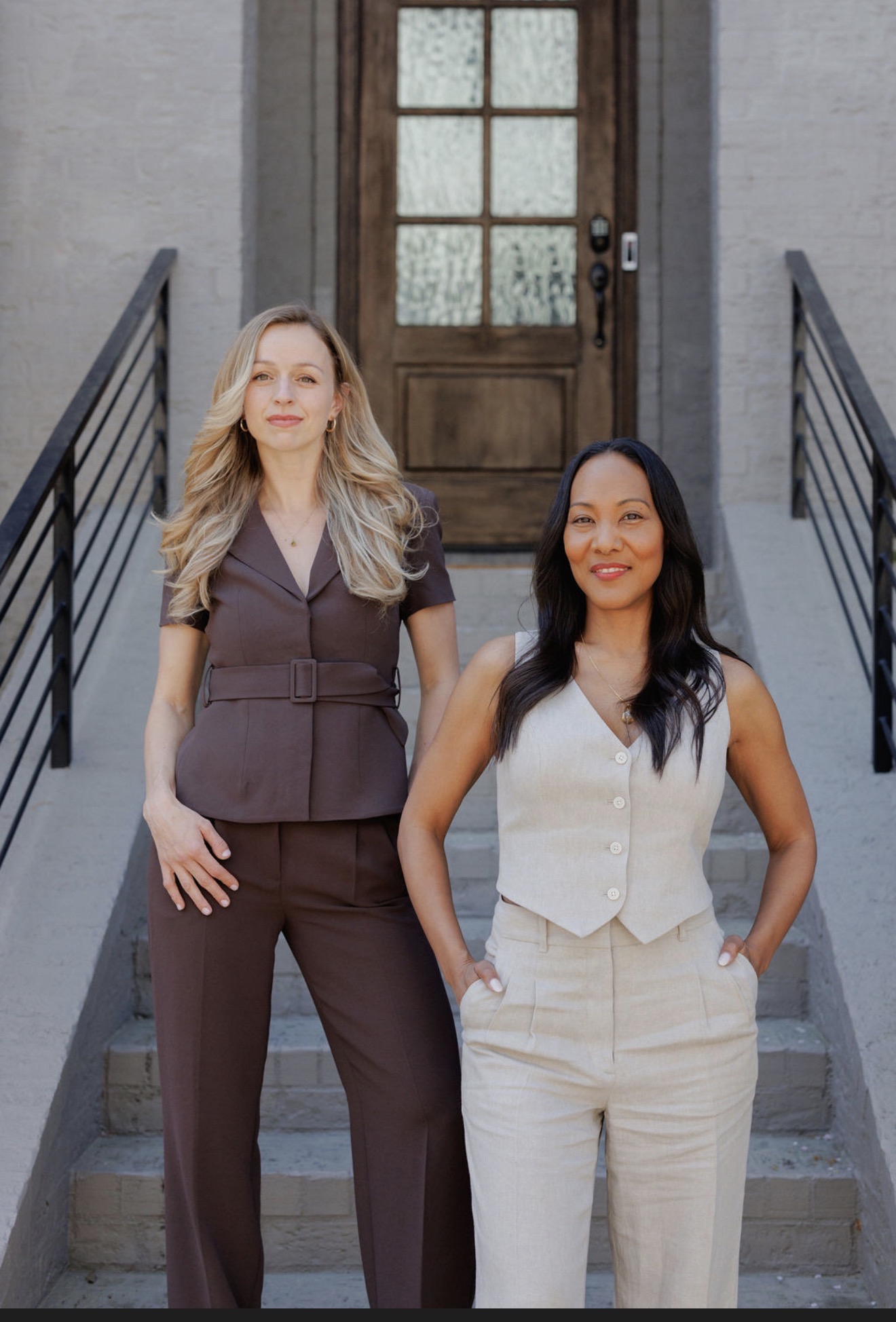 Two women stand on stairs in front of a door with glass panes, smiling and posing for the photo.