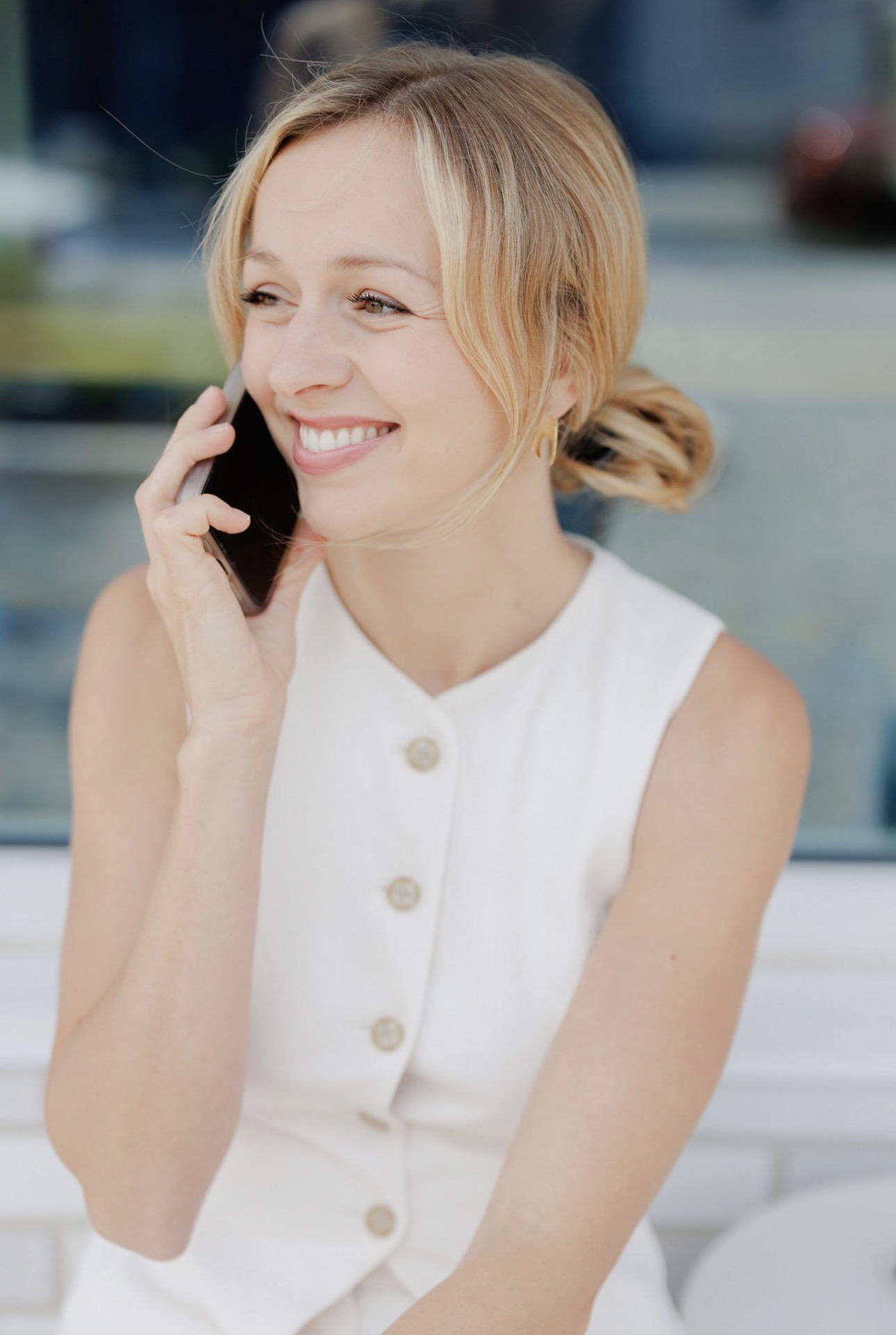 Woman with blonde hair smiling and talking on a phone outdoors, wearing a white sleeveless top.