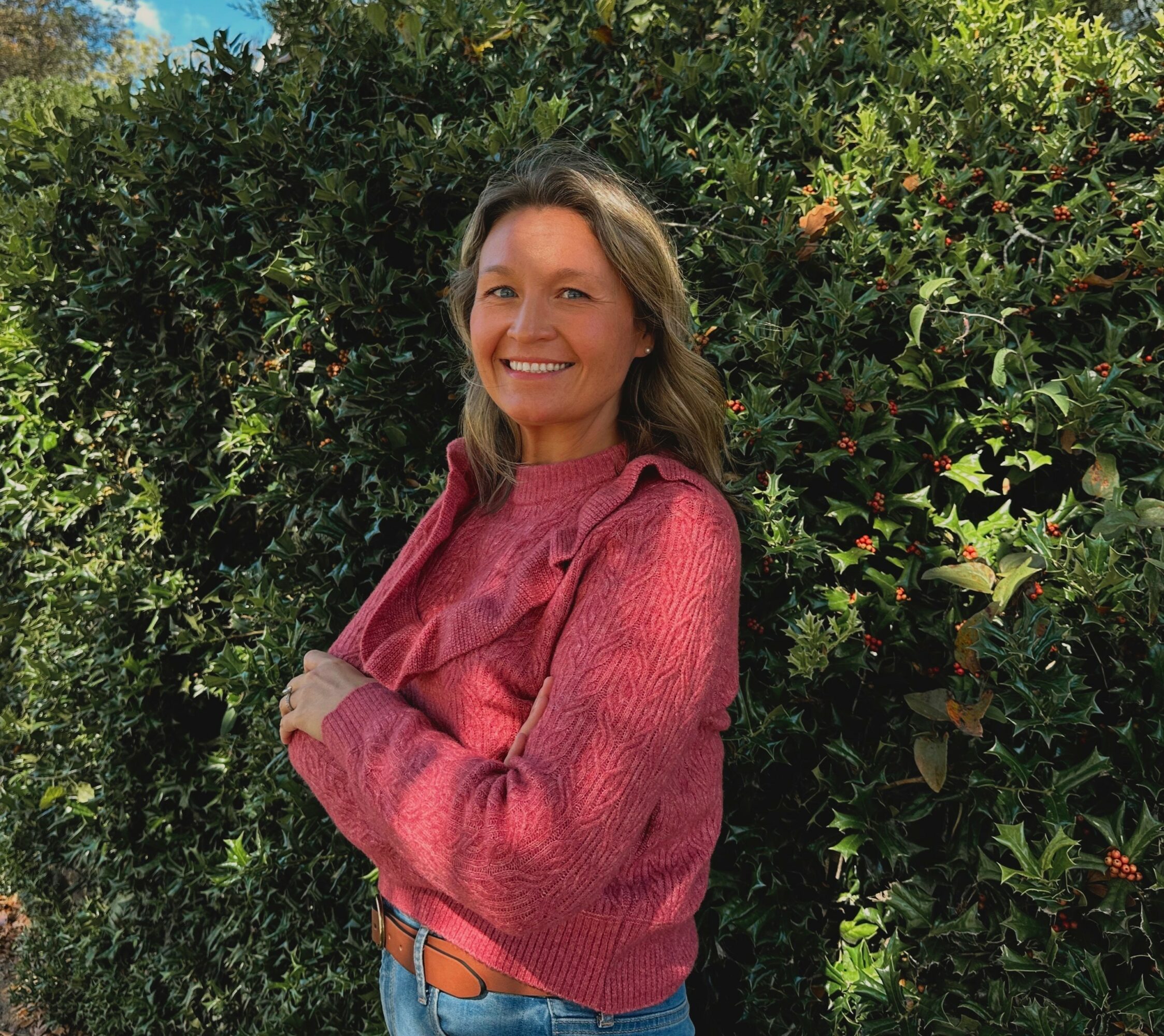 Woman with brown hair smiling, wearing a pink sweater, standing outdoors in front of green bushes.