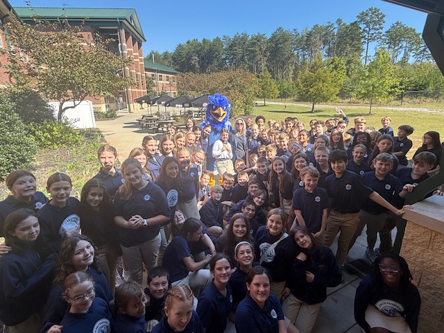 Large group of students gathered outdoors with a person in a blue mascot costume, school building and trees in background.